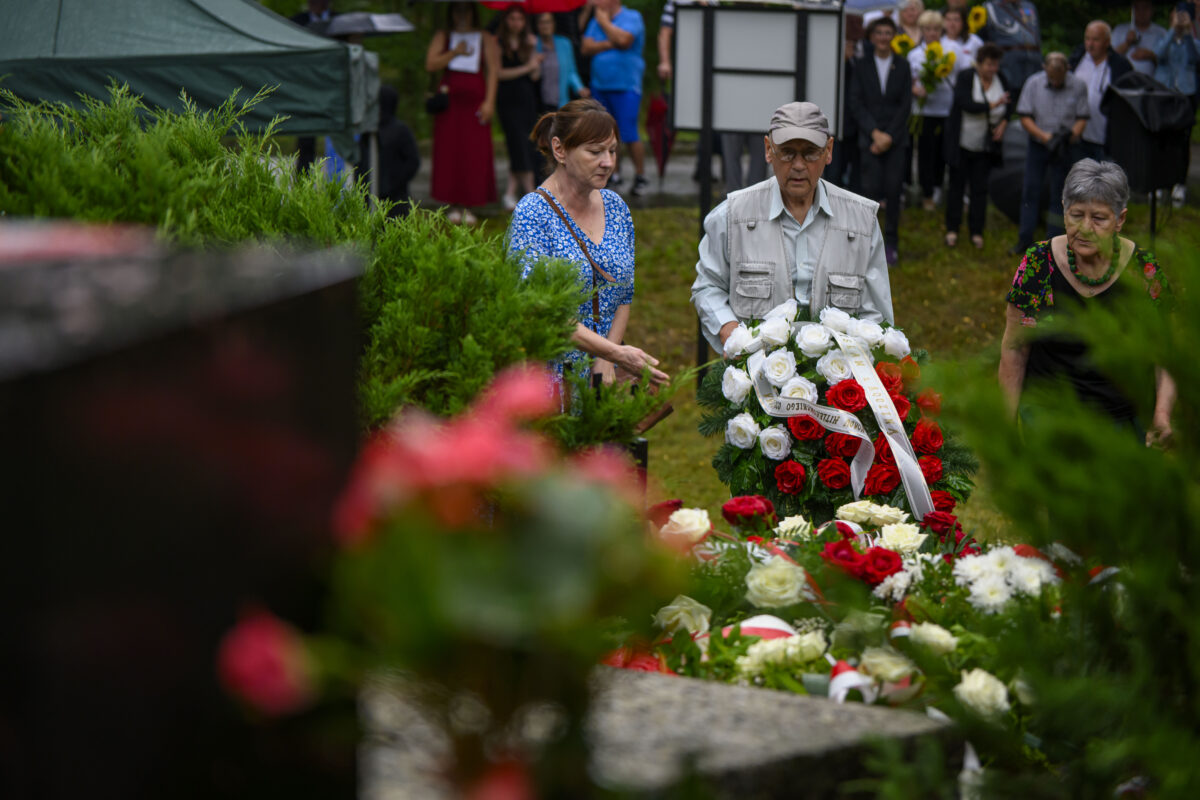 Trzy osoby składają wieniec z czerwonych i białych róż na cmentarzu. Tło jest zielone, z grupą ludzi w oddali. Scena pełna kwiatów.