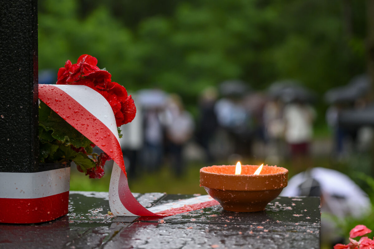 Bukiet czerwonych kwiatów z biało-czerwoną wstążką leży obok płonącego znicza. Tło zamglone, zielone drzewa i ludzie. Pada deszcz.