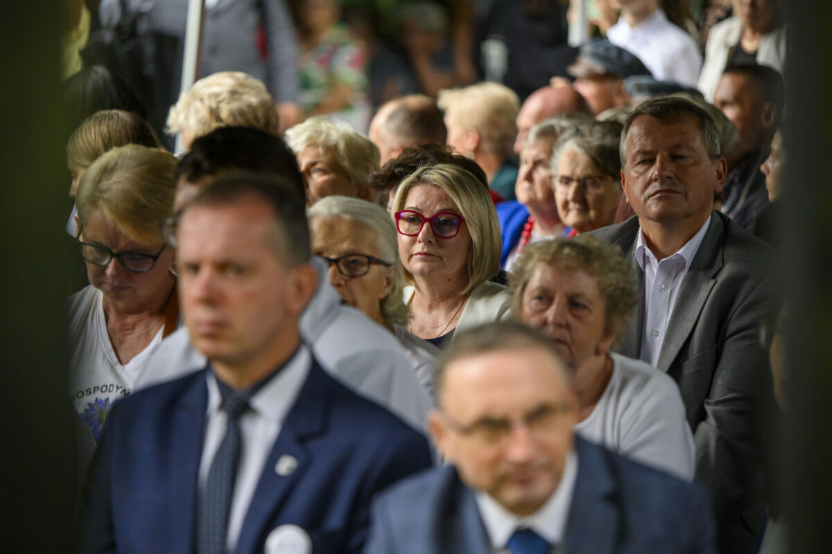Grupa ludzi siedzi w rzędach; kobieta z różowymi okularami na środku. Tło jest rozmyte, dominuje kolor zielony i biały.
