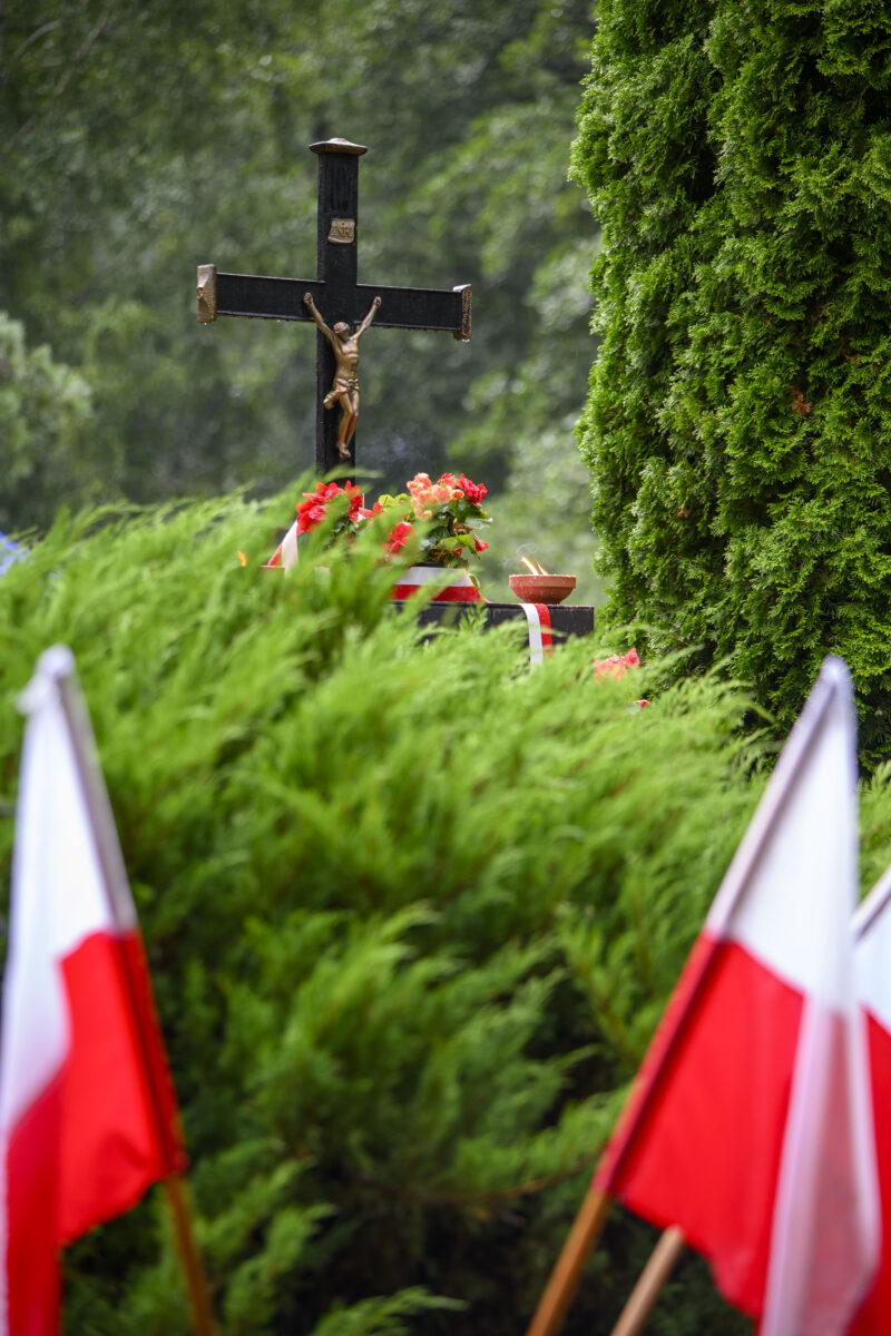 Czarne krzyż z Jezusem na tle zielonych drzew, otoczony czerwonymi kwiatami i polskimi flagami.