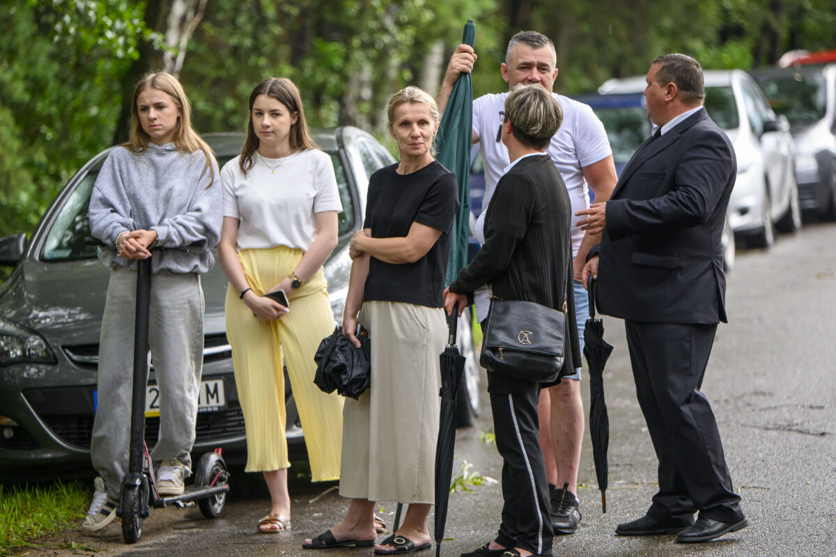 Grupa ludzi stoi na ulicy z zielenią w tle. Jedna osoba trzyma hulajnogę, inna ma czarną parasolkę. Kobieta w czarnym swetrze nosi torbę na ramieniu.