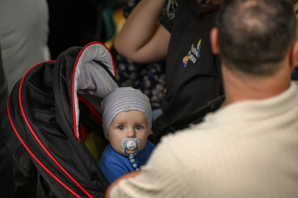 Na zdjęciu widoczny jest niemowlak w czapce, z smoczkiem, siedzący w wózku o ciemnym kolorze z czerwonymi akcentami. Obok dziecka znajduje się dorosły. Tło jest rozmyte, ciemne.