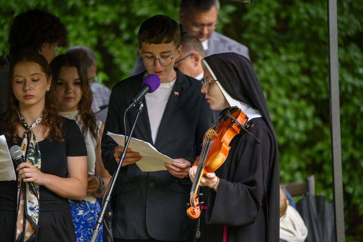 Grupa ludzi stoi na scenie z mikrofonami, jedna osoba trzyma skrzypce. W tle zielone liście. Kolorystyka stonowana.