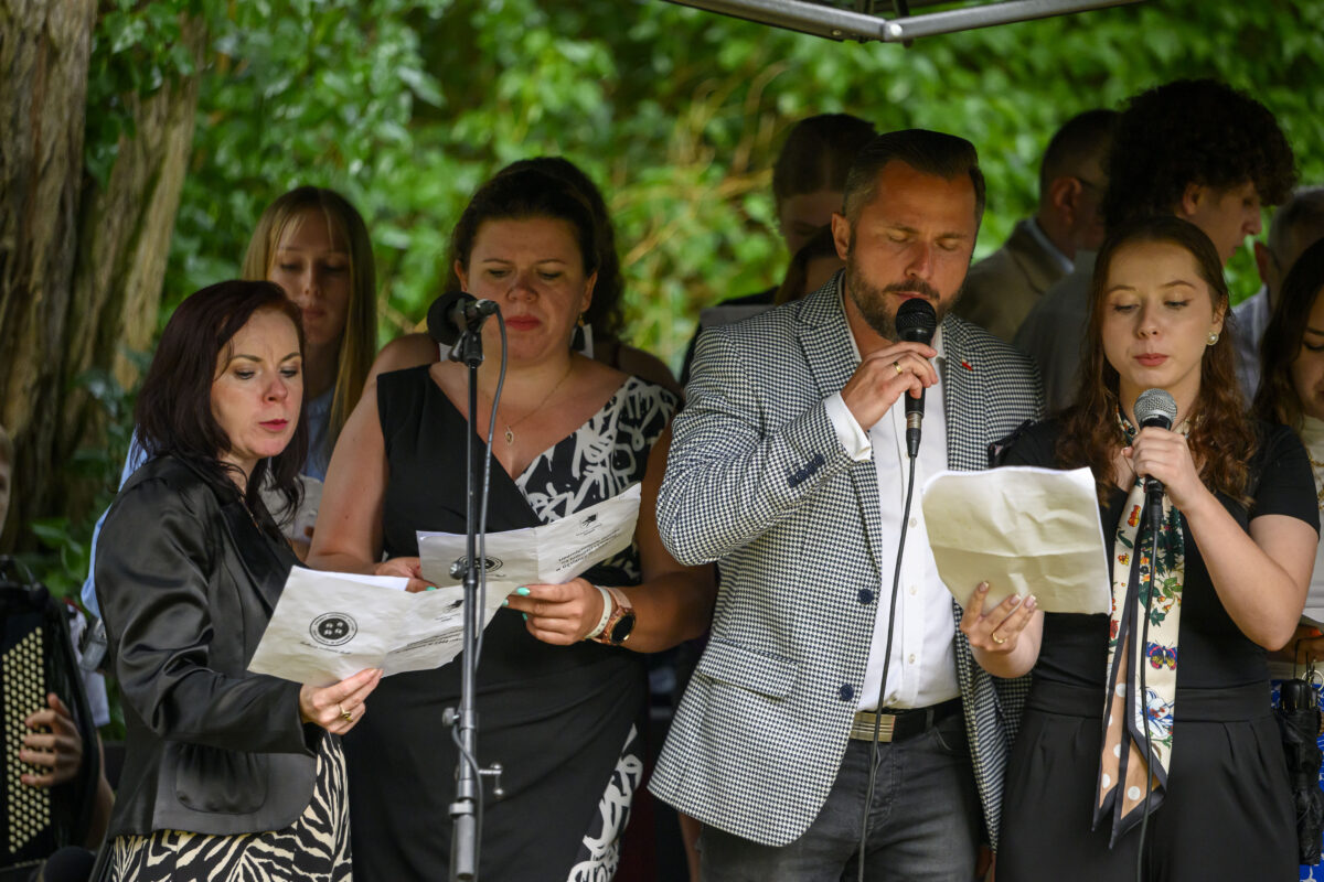 Grupa osób śpiewa z nutami w rękach. Są na zewnątrz, otoczeni zielonymi drzewami. Mają czarno-białe ubrania.