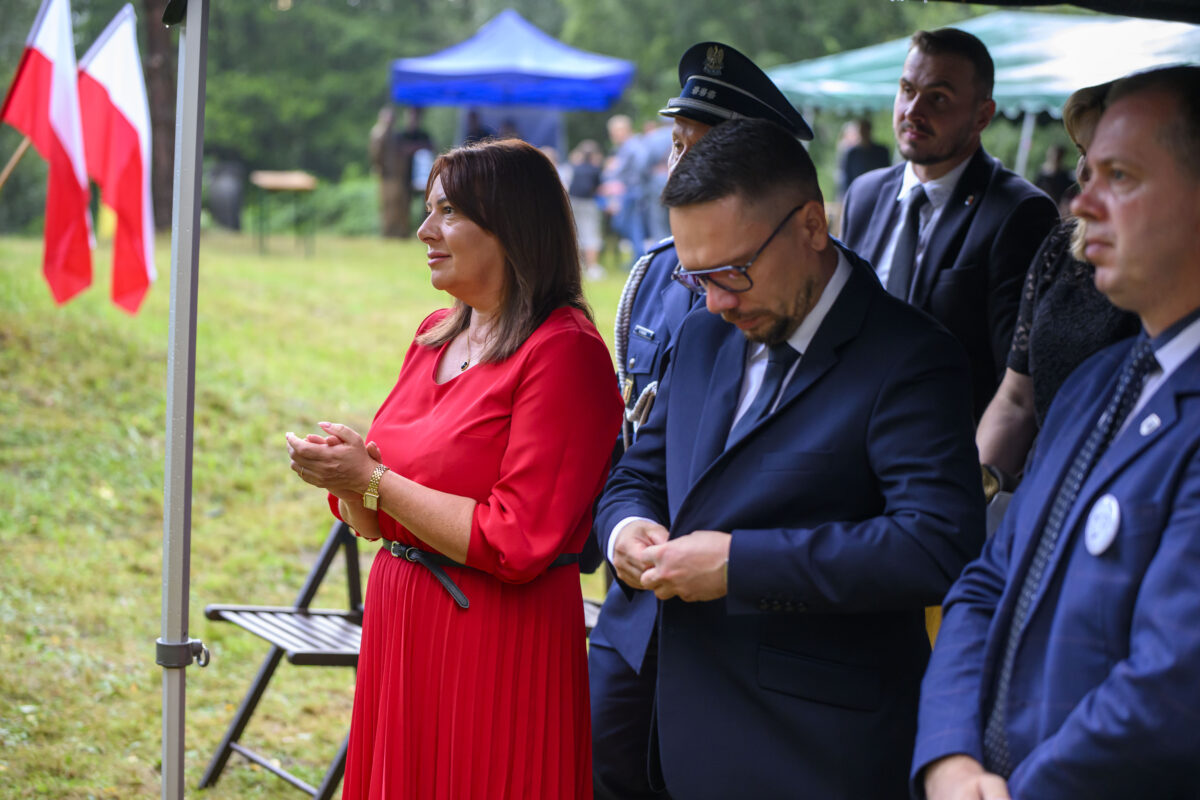 Grupa ludzi na zewnątrz, kobieta w czerwonej sukience, mężczyźni w garniturach. W tle zielona trawa i polskie flagi.