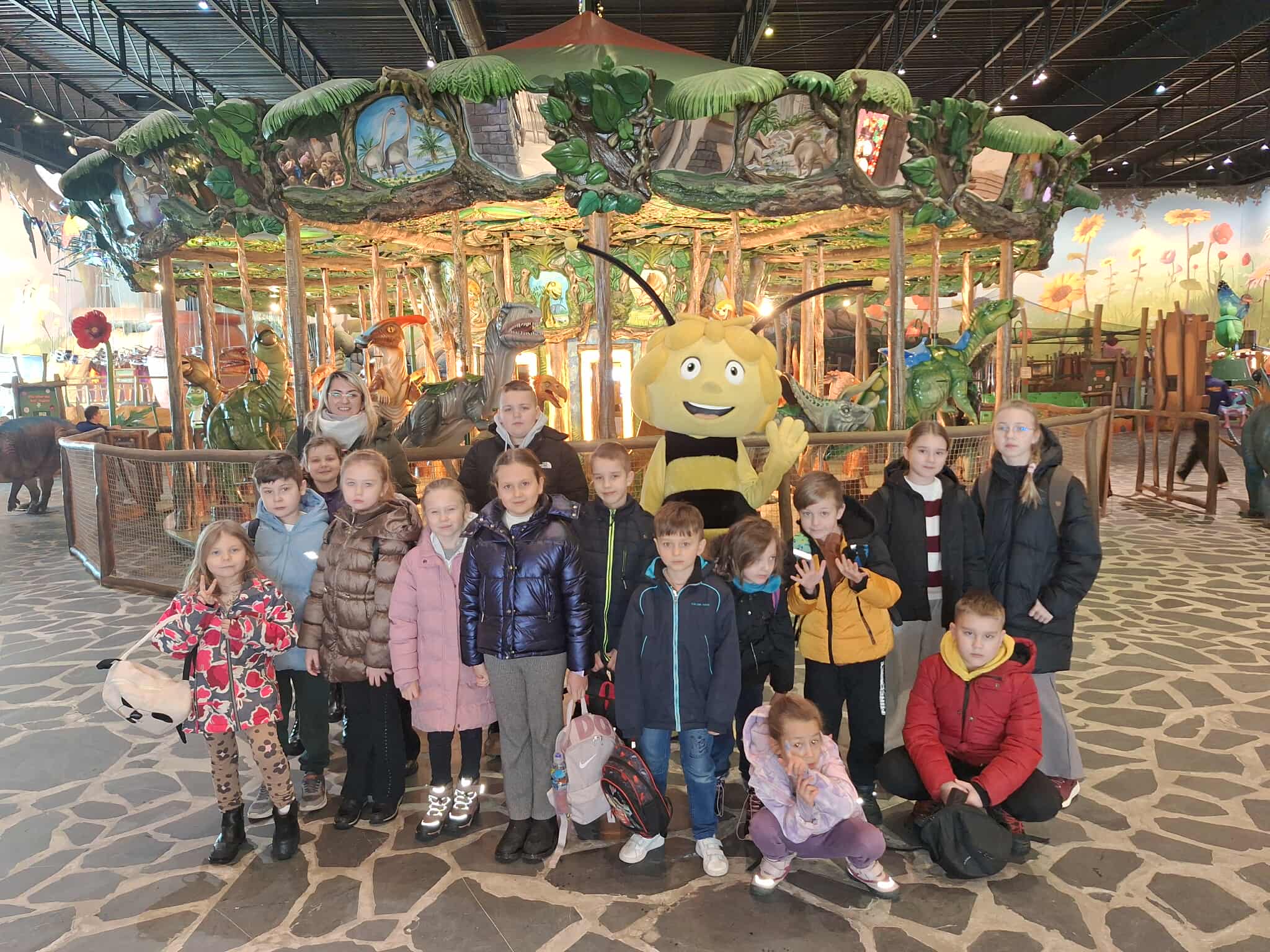 Grupa dzieci pozuje przed kolorową karuzelą z figurami dinozaurów. W tle dominuje bujna, zielona sceneria. Maskotka pszczoły towarzyszy dzieciom na pierwszym planie.