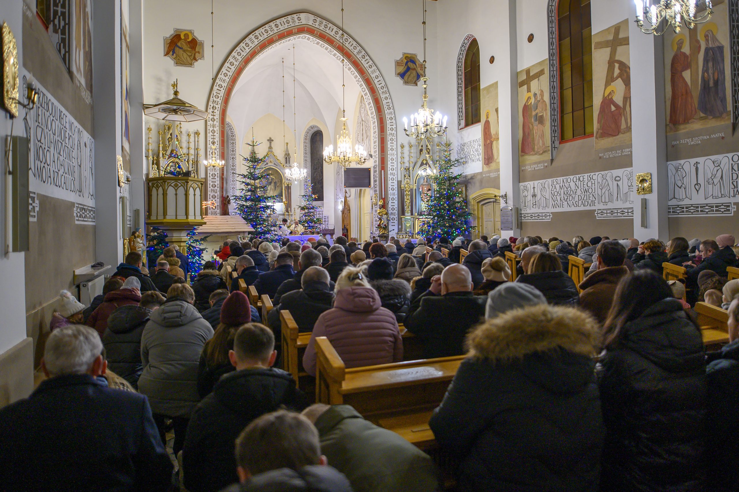 Wnętrze kościoła z tłumem wiernych. Bogato zdobione ściany, choinki i żyrandole. Atmosfera świąteczna, kolory złote i białe.