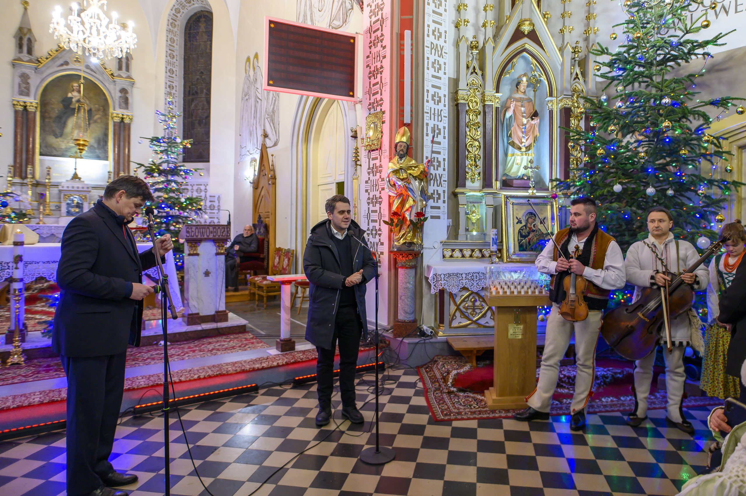 Kościół z dekoracjami i muzykami w ludowych strojach. Jasne ściany, kolorowe oświetlenie i choinka. Dwie osoby przy mikrofonach.