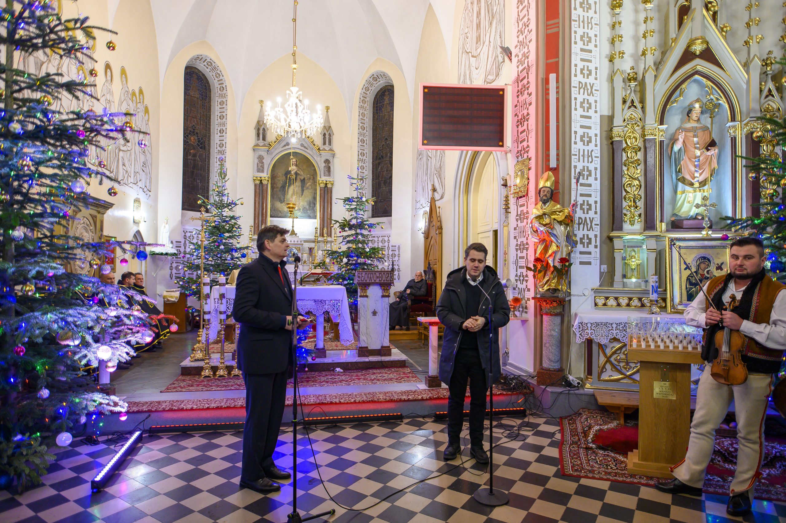 Wnętrze kościoła z dekoracjami świątecznymi, choinkami i ołtarzem. Na pierwszym planie stoją trzy osoby przy mikrofonach. Podłoga w czarno-białą szachownicę.
