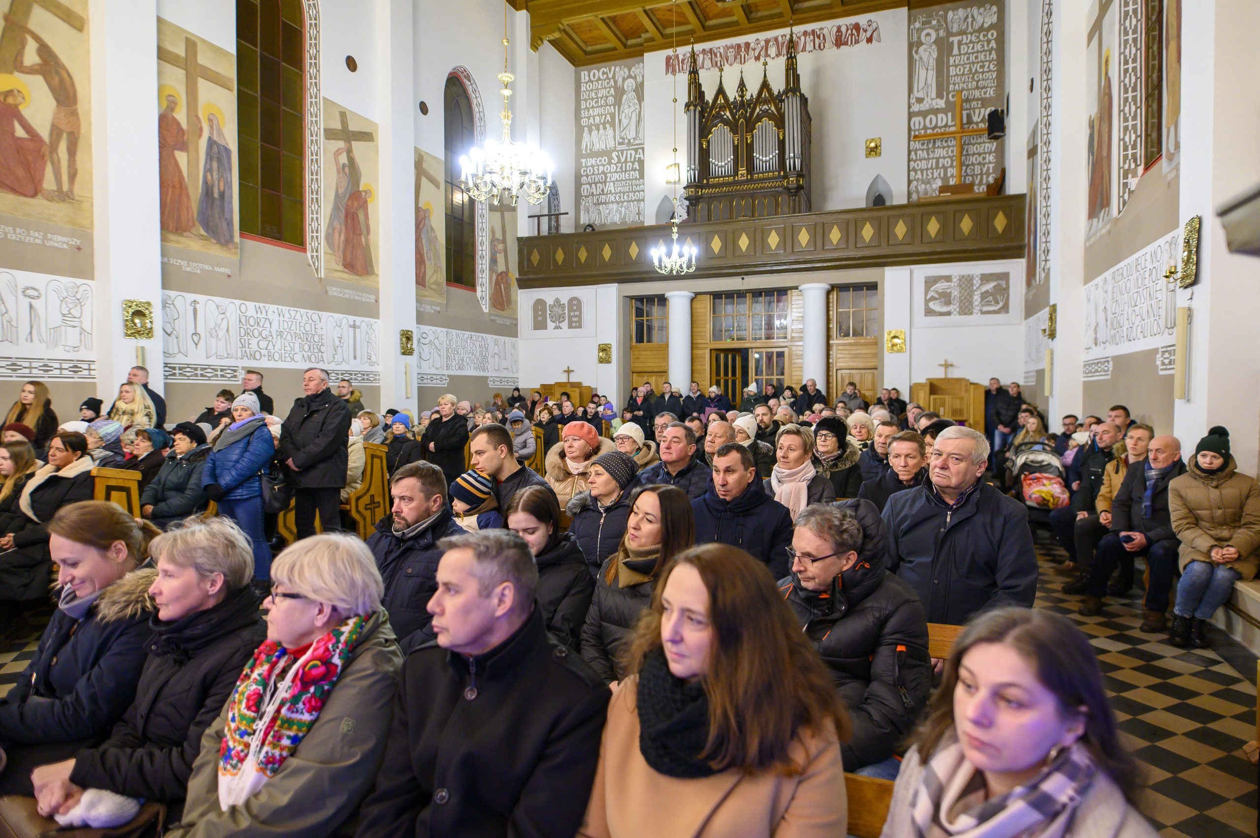 Grupa ludzi siedzi w kościele z malowidłami na ścianach. Jasne wnętrze z żyrandolami i drewnianym ołtarzem. Tło z białymi i brązowymi elementami.