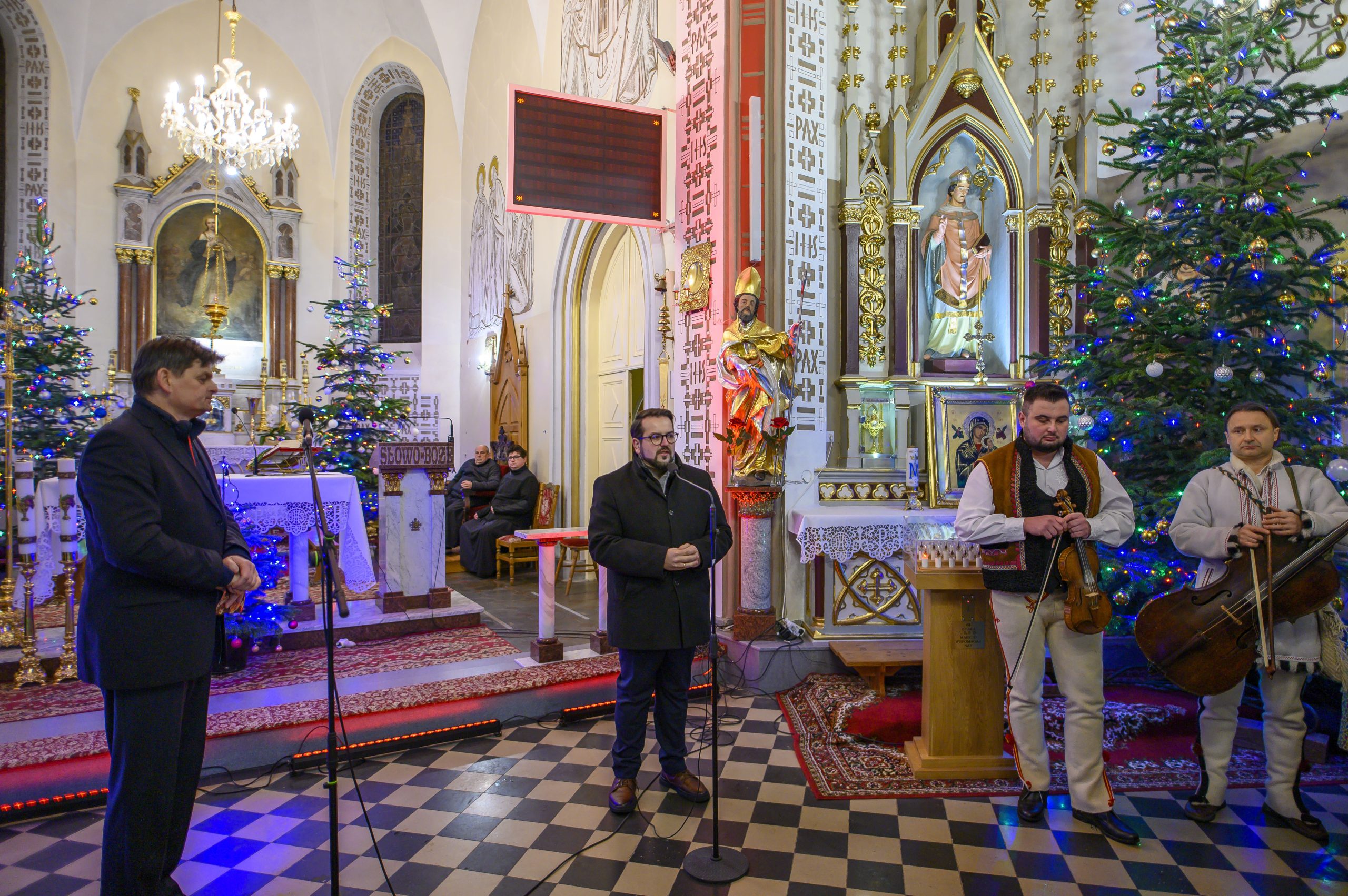 Grupa mężczyzn w kościele na tle ołtarza i choinek. Ornaments stylu gotyckiego i jasne światła. Na podłodze czarno-biała szachownica.
