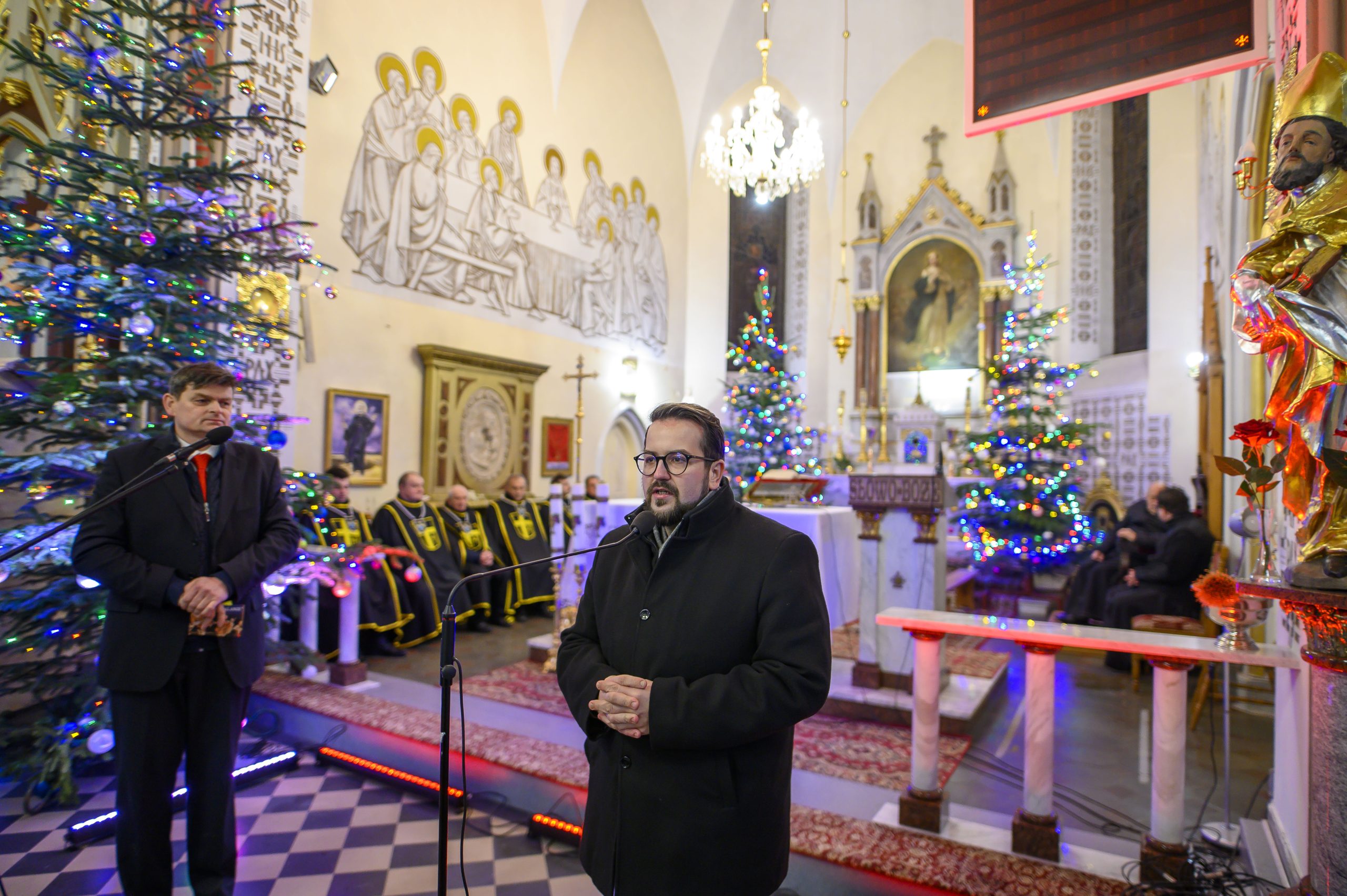 Mężczyzna w czarnym płaszczu przemawia przy mikrofonie w kościele. Tło zdobią choinki z kolorowymi lampkami i freski. Wnętrze ma jasne ściany i dekorowane ołtarze.