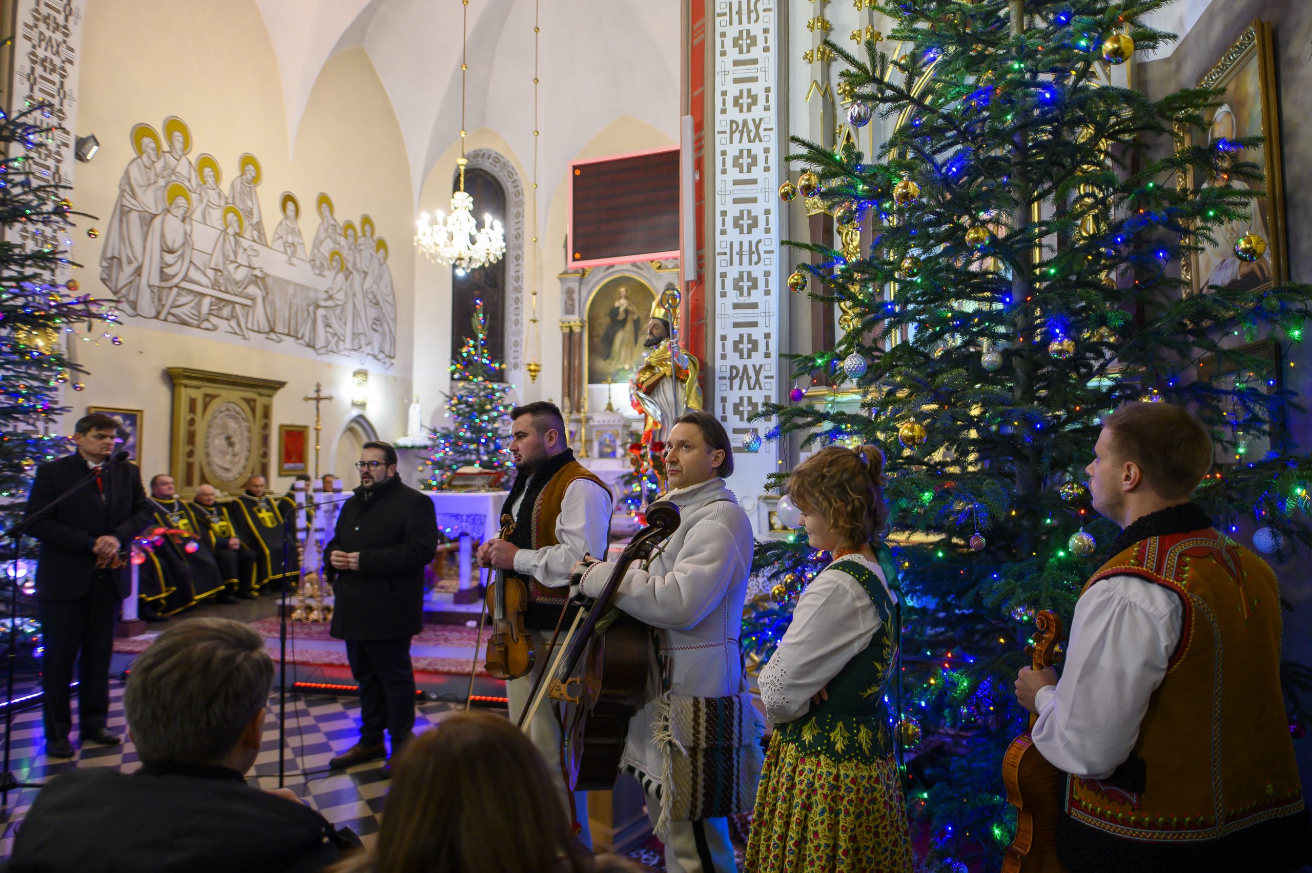 Ludzie w strojach ludowych grają na instrumentach w kościele. W tle choinki z kolorowymi światełkami. Atmosfera świąteczna.