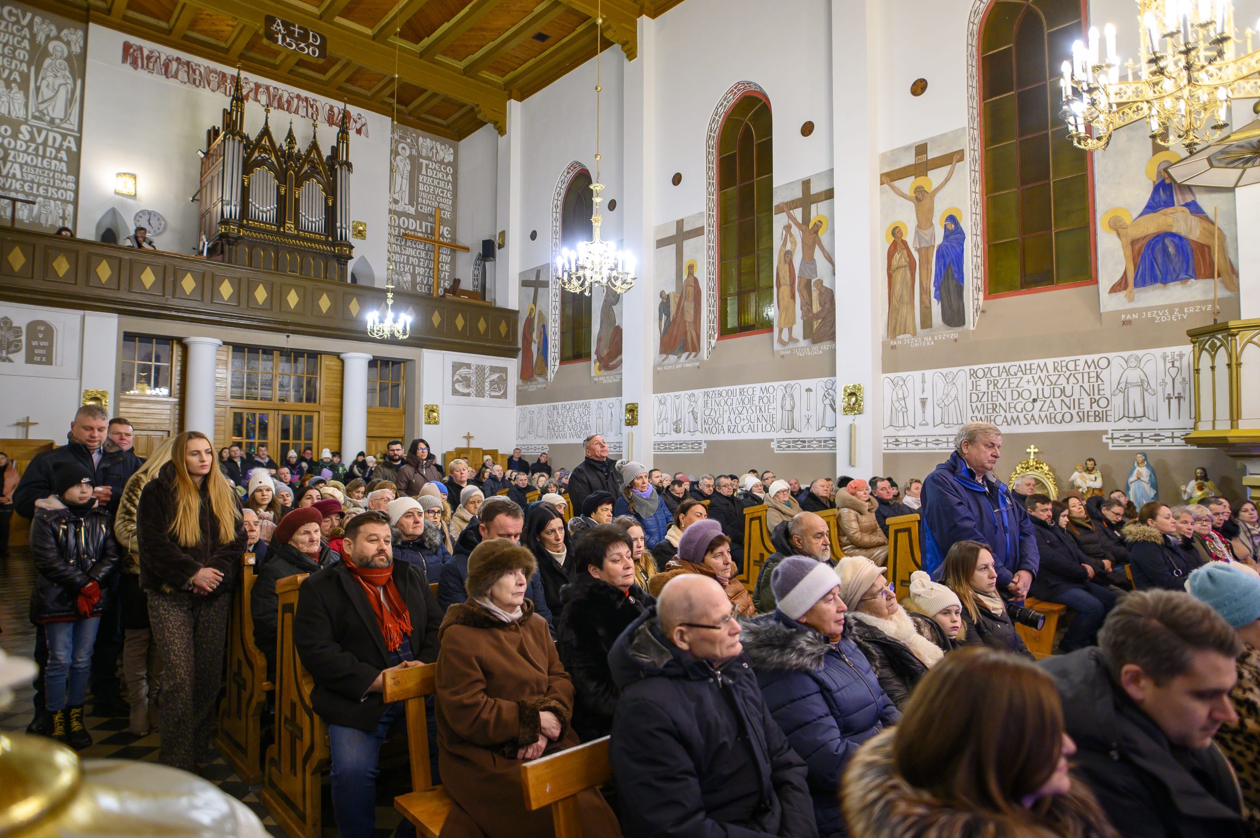 Na zdjęciu widać wnętrze kościoła z dużą liczbą osób siedzących na ławkach. Ściany są ozdobione malowidłami i napisami. Wiszące żyrandole oświetlają przestrzeń ciepłym światłem.