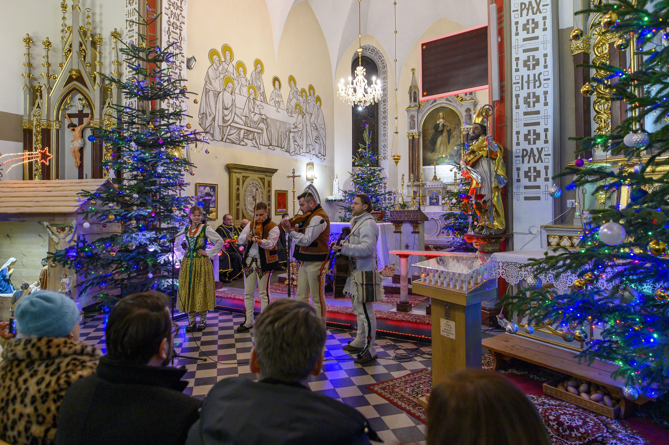 Grupa muzyków w strojach ludowych gra w kościele, otoczona ozdobnymi choinkami. Ściany zdobią freski, a wnętrze jest bogato oświetlone. Widownia siedzi na czarno-białej podłodze.
