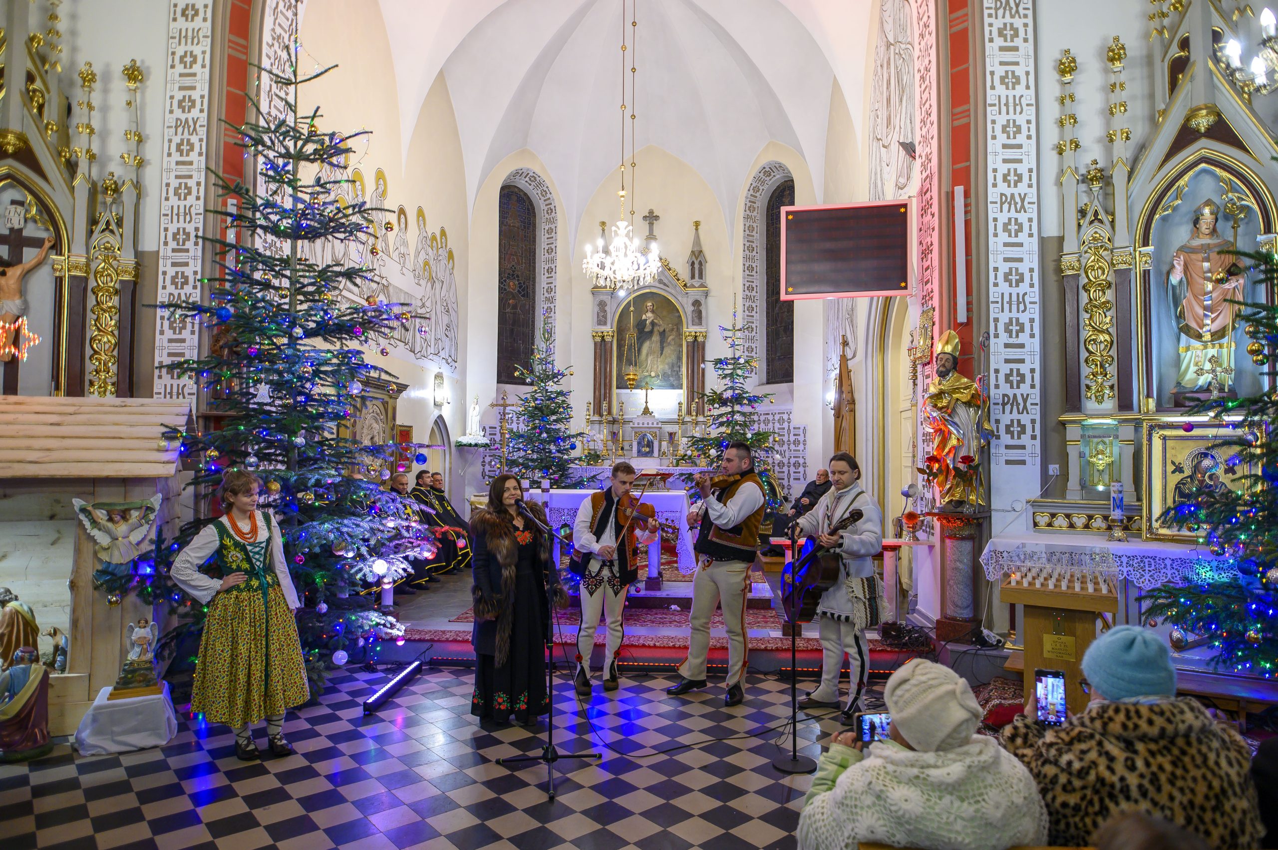 Grupa muzyków w ludowych strojach gra w kościele. W tle choinki, dekoracje i złote detale na ścianach. Publiczność siedzi i robi zdjęcia.