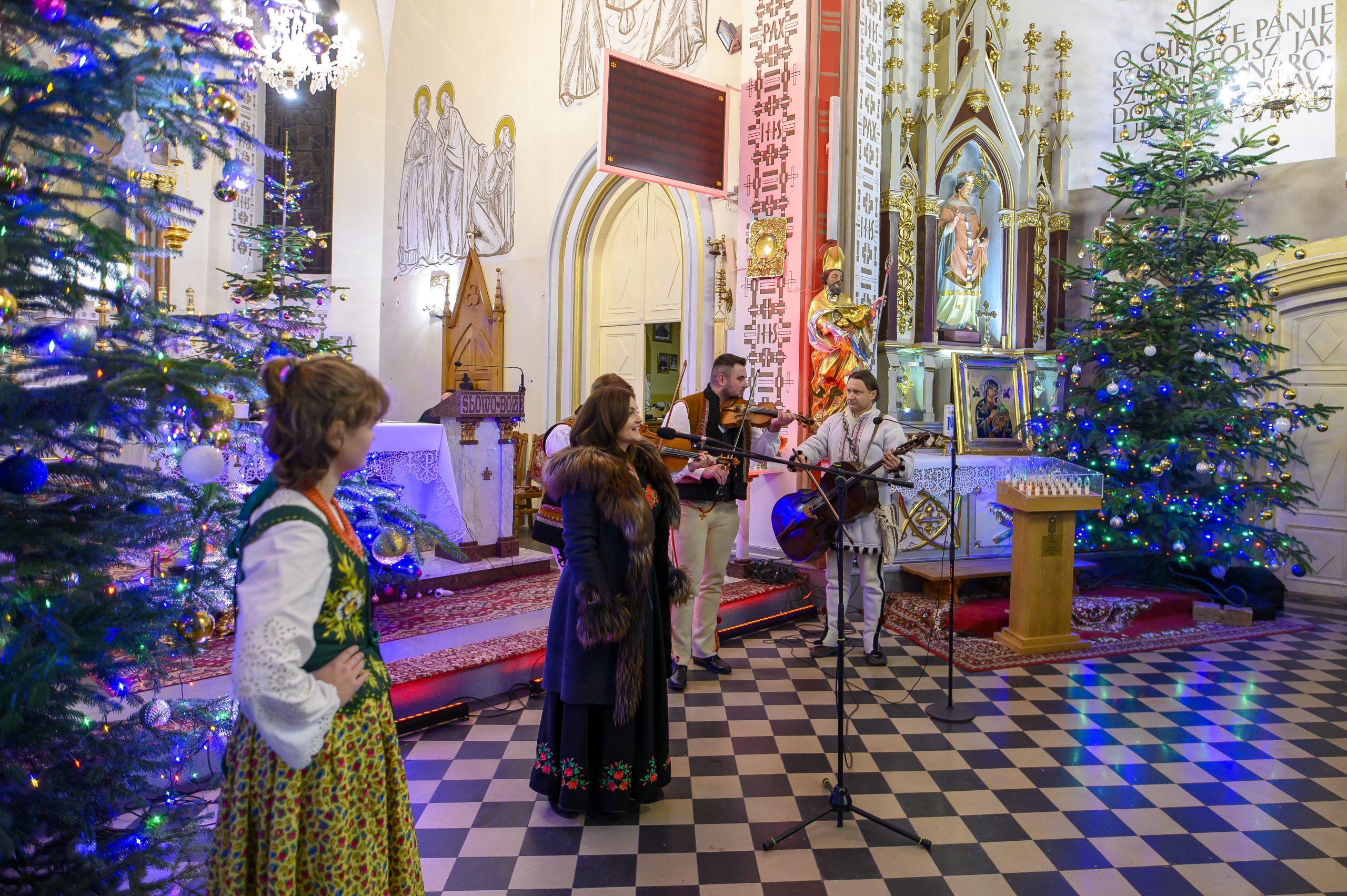 Grupa muzyków w strojach ludowych gra w kościele, ozdobionym kolorowymi światełkami i choinkami. Wnętrze utrzymane jest w jasnych barwach ze złotymi zdobieniami. Na podłodze czarno-biała szachownica.
