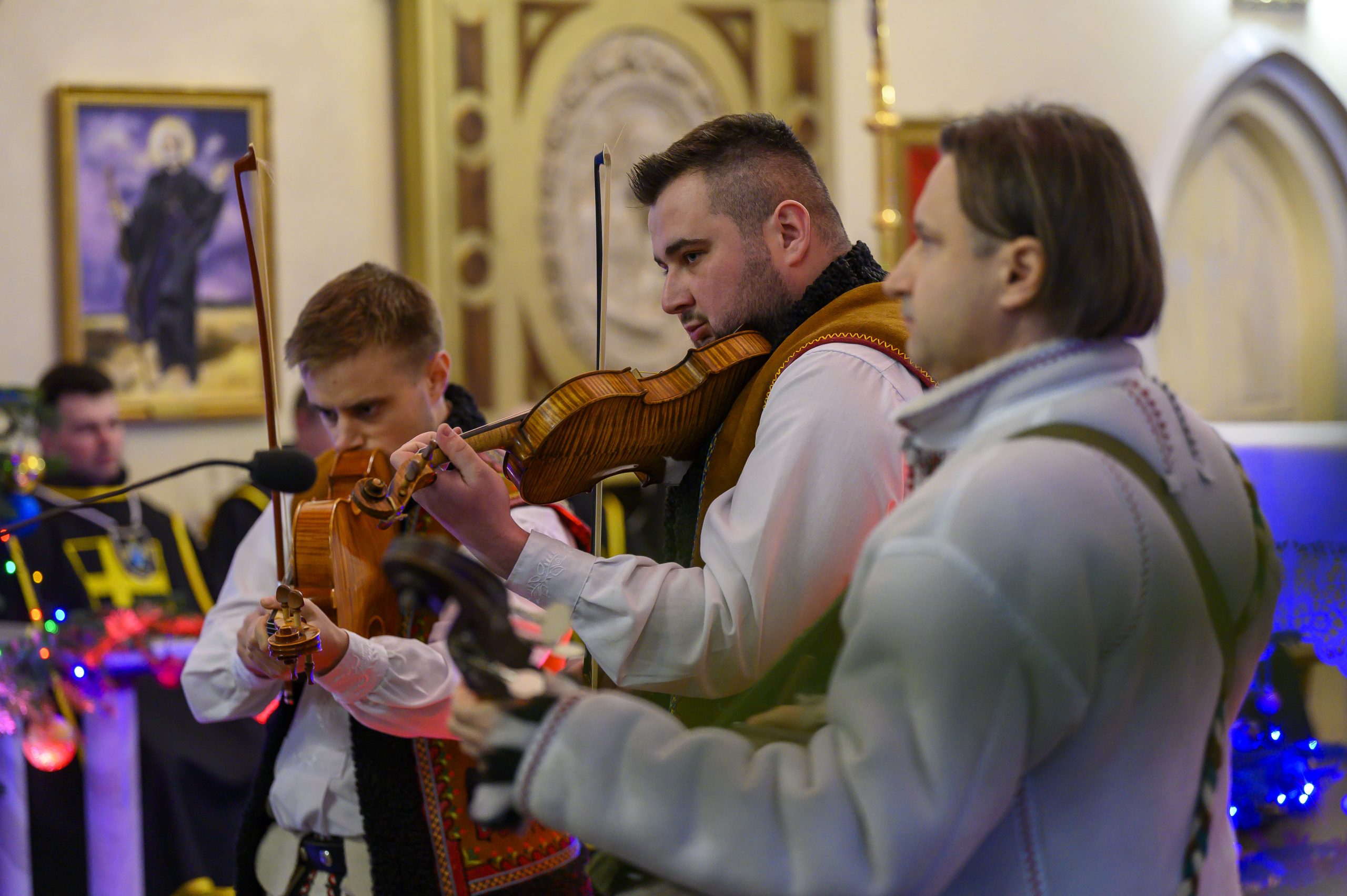 Muzycy w tradycyjnych strojach grają na skrzypcach w kościele. Dekoracje w tle są kolorowe. Ściany i malowidła w odcieniach beżu.