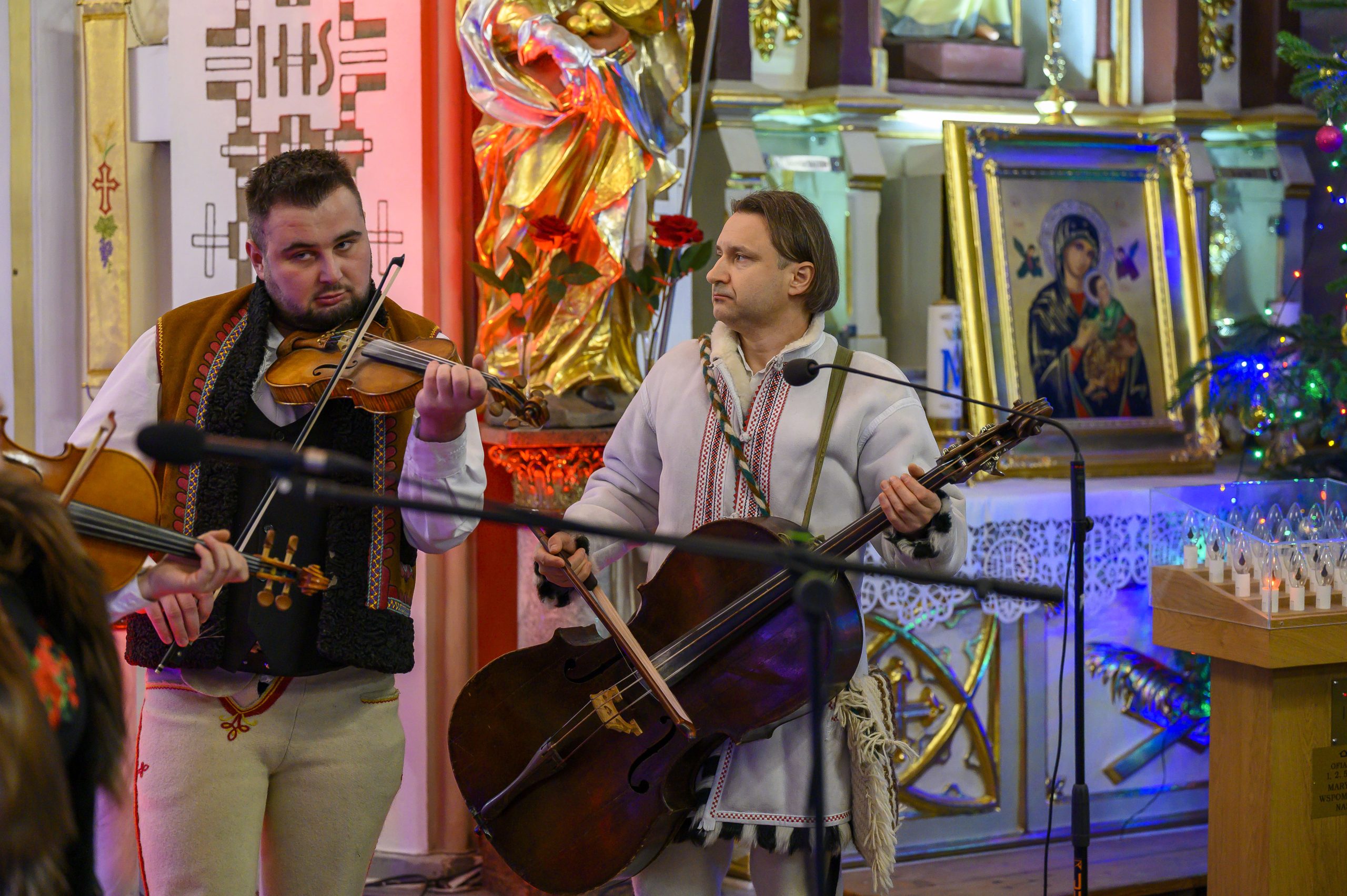 Dwóch mężczyzn gra na instrumentach smyczkowych w kolorowym wnętrzu kościoła. Tło ozdobione złotą rzeźbą i ikoną Matki Boskiej. Oświetlenie w ciepłych tonacjach.