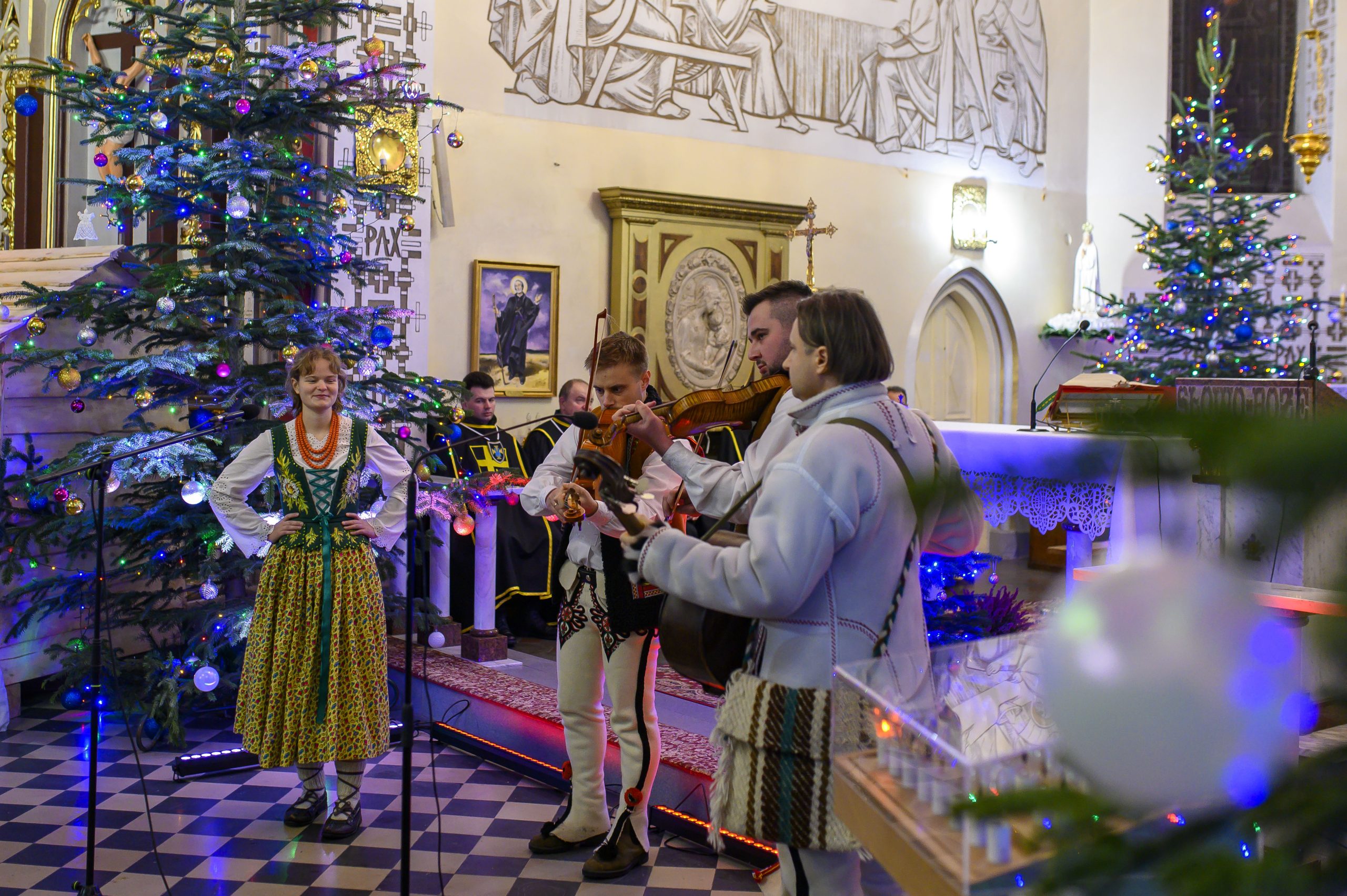 Grupa ludzi w tradycyjnych strojach góralskich gra muzykę w kościele ozdobionym choinkami. W tle ściany z freskami. Kolory: zieleń, biel, brąz.