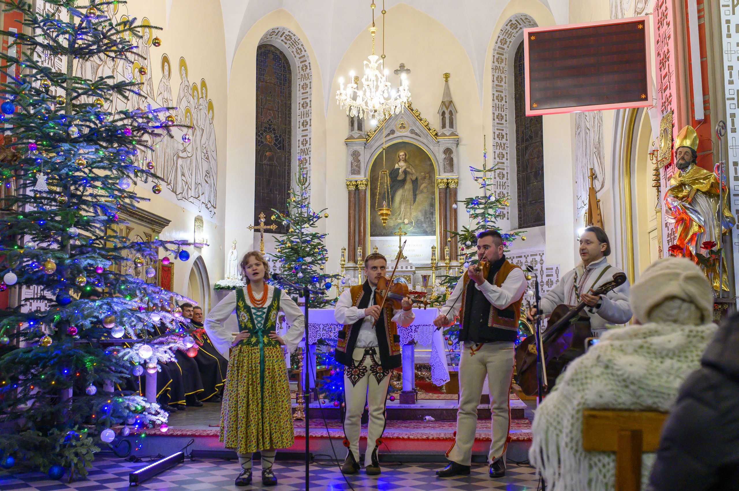 Grupa muzyków w tradycyjnych strojach występuje w udekorowanym świątecznie kościele. Ściany zdobią freski i witraże, a wnętrze oświetla żyrandol. Choinki z kolorowymi lampkami tworzą świąteczny nastrój