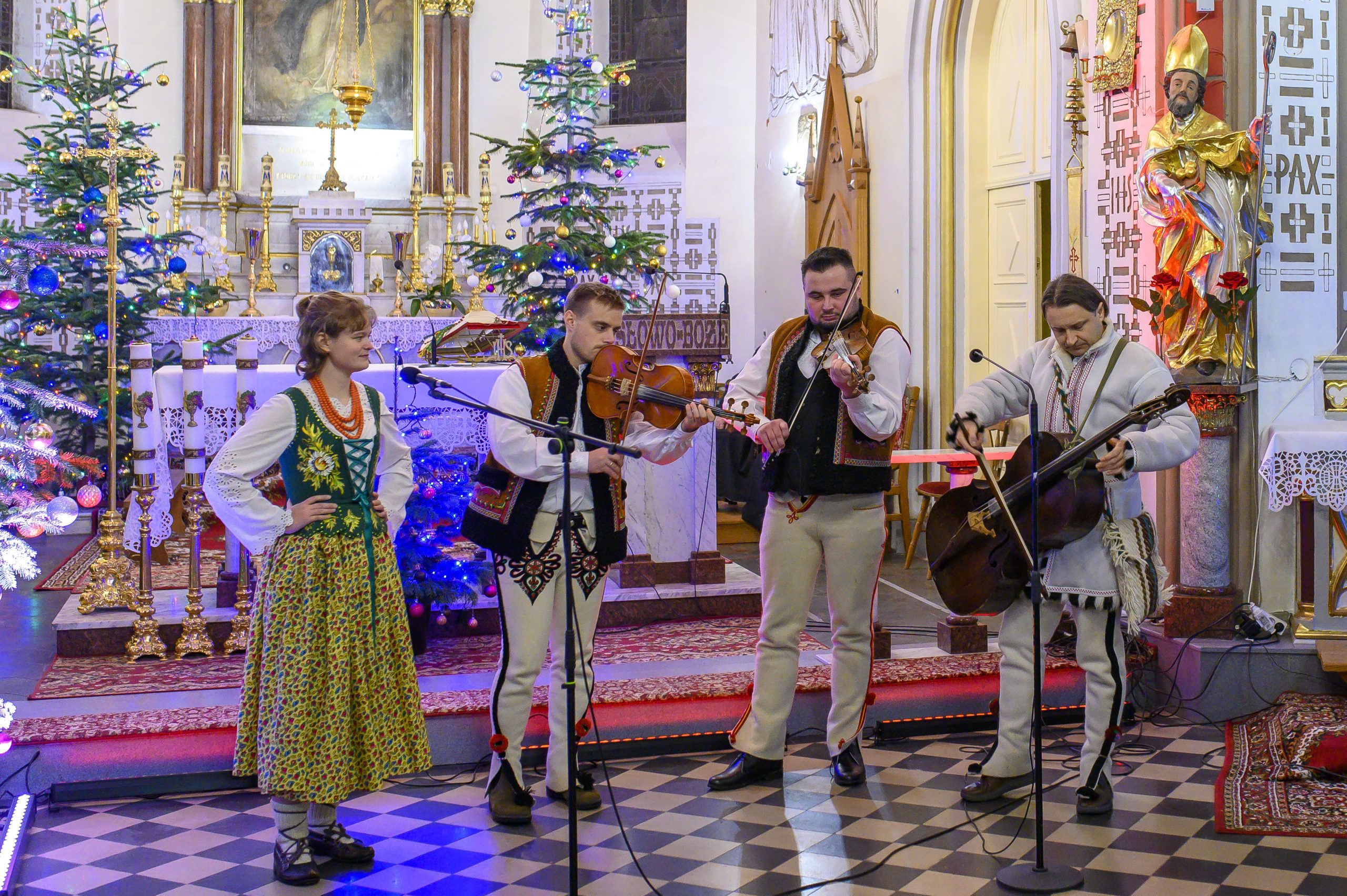 Grupa muzyków w strojach ludowych gra w kościele ozdobionym choinkami. Kolorowe światełka i czerwony dywan tworzą tło. Postacie są otoczone świątecznymi dekoracjami.