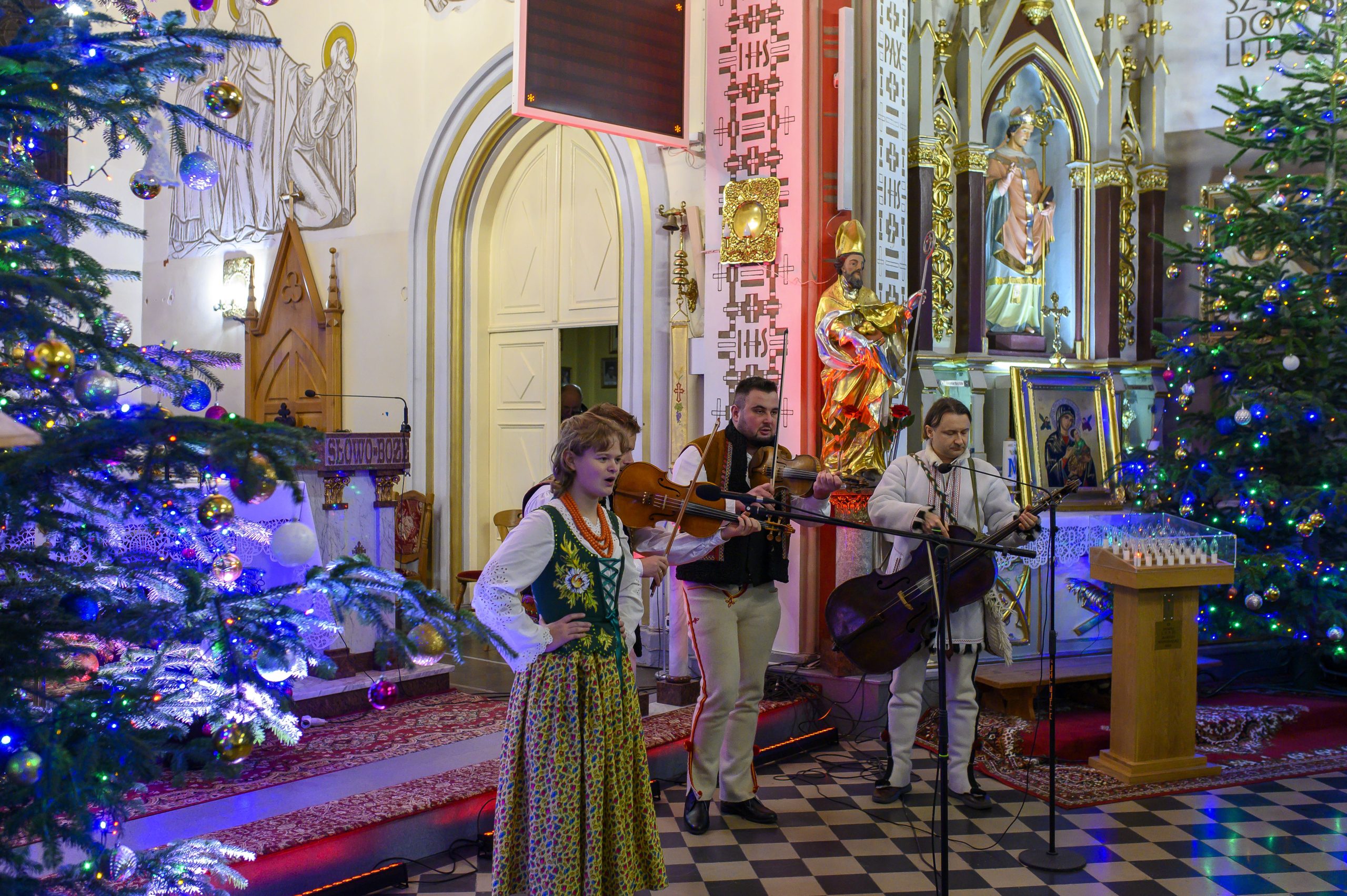 Grupa muzyków gra przed ołtarzem w kościele, ubranych w tradycyjne stroje. Obok dekoracje świąteczne z choinkami i lampkami. W tle złote ornamenty i ikony.