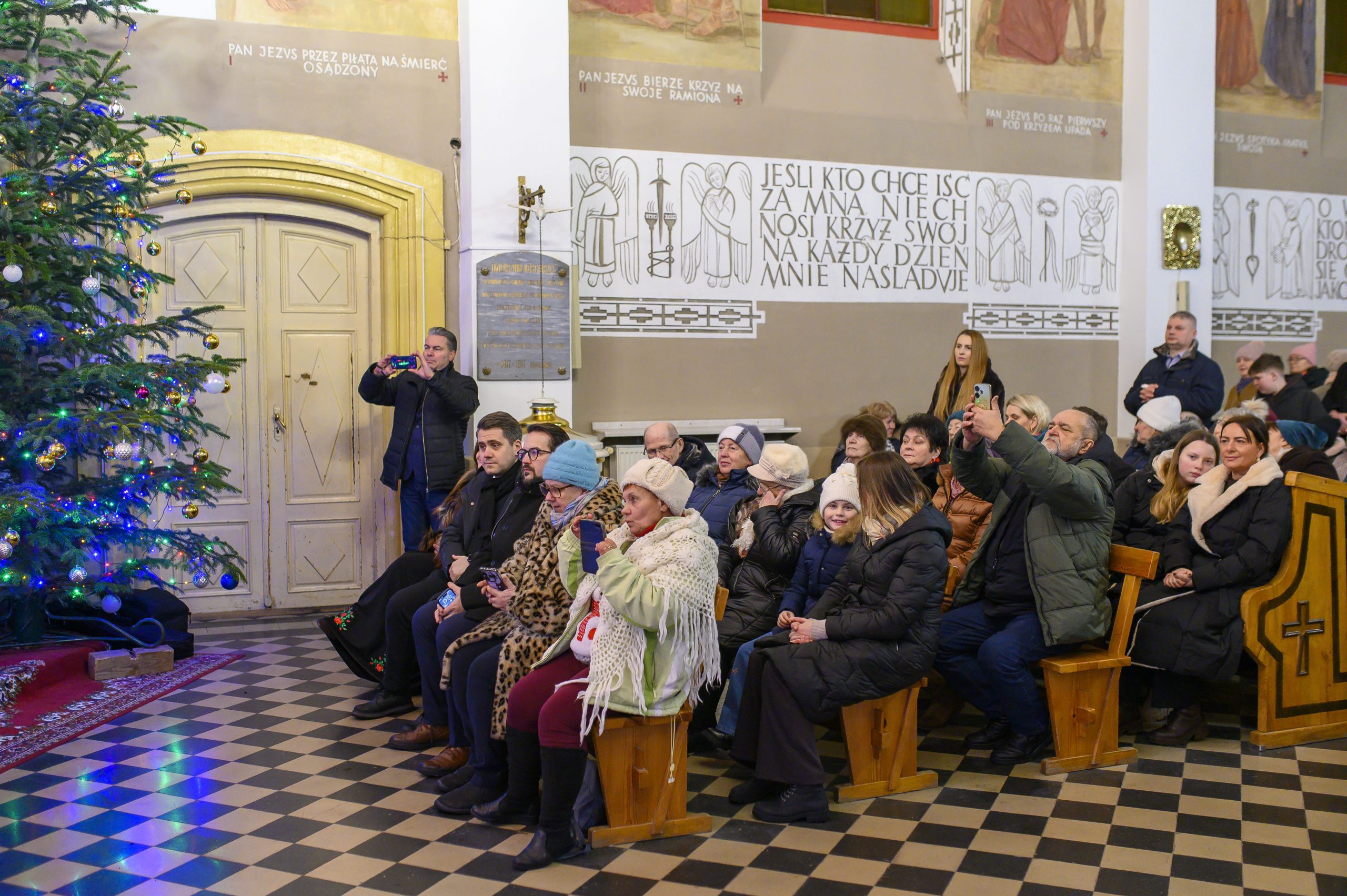 Grupa ludzi siedzi w drewnianych ławkach kościoła, obok choinka z ozdobami. Na tle ściana z religijnymi inskrypcjami. Granie światłem nadaje ciepły nastrój.