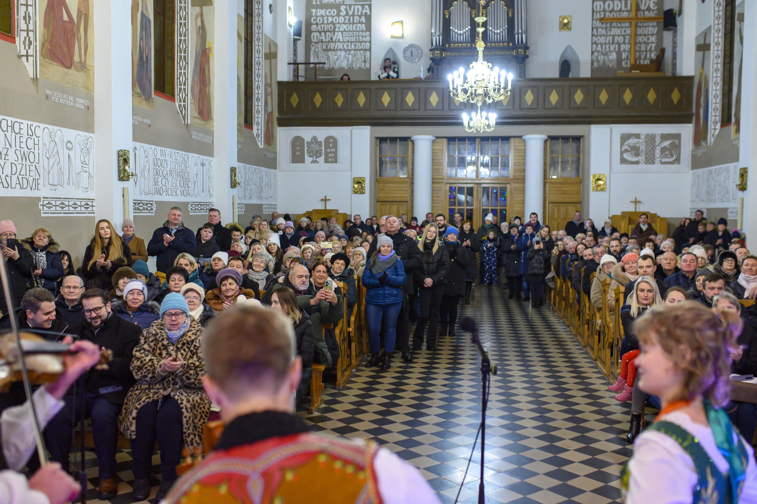 Ludzie zgromadzeni w kościele z jasnym wnętrzem, ozdobnymi freskami i żyrandolem. Na pierwszym planie muzycy w tradycyjnych strojach. Atmosfera jest radosna.