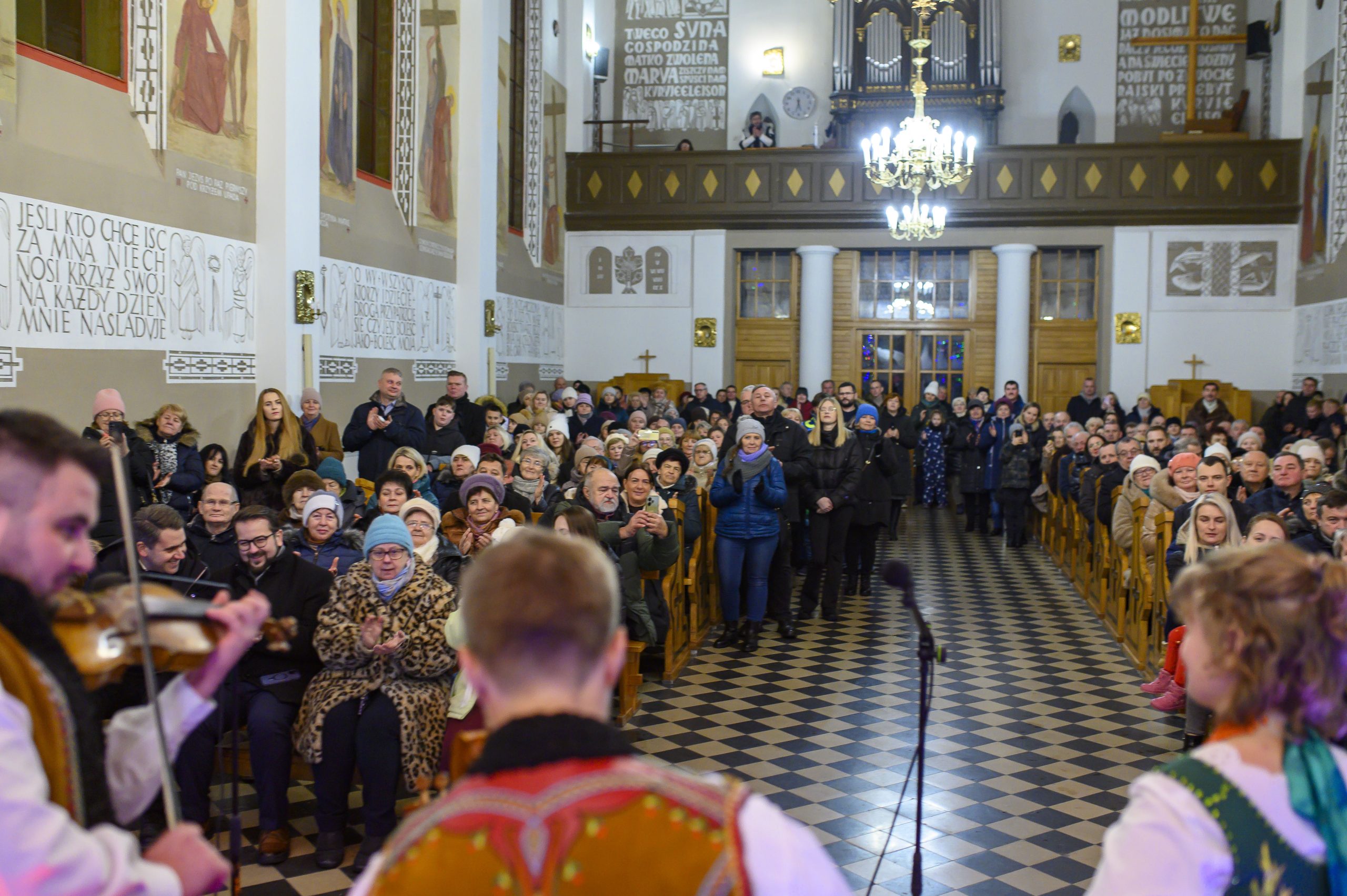 Grupa ludzi w kościele o jasnym wnętrzu, błyszczący żyrandol i muzycy w tradycyjnych strojach na pierwszym planie.