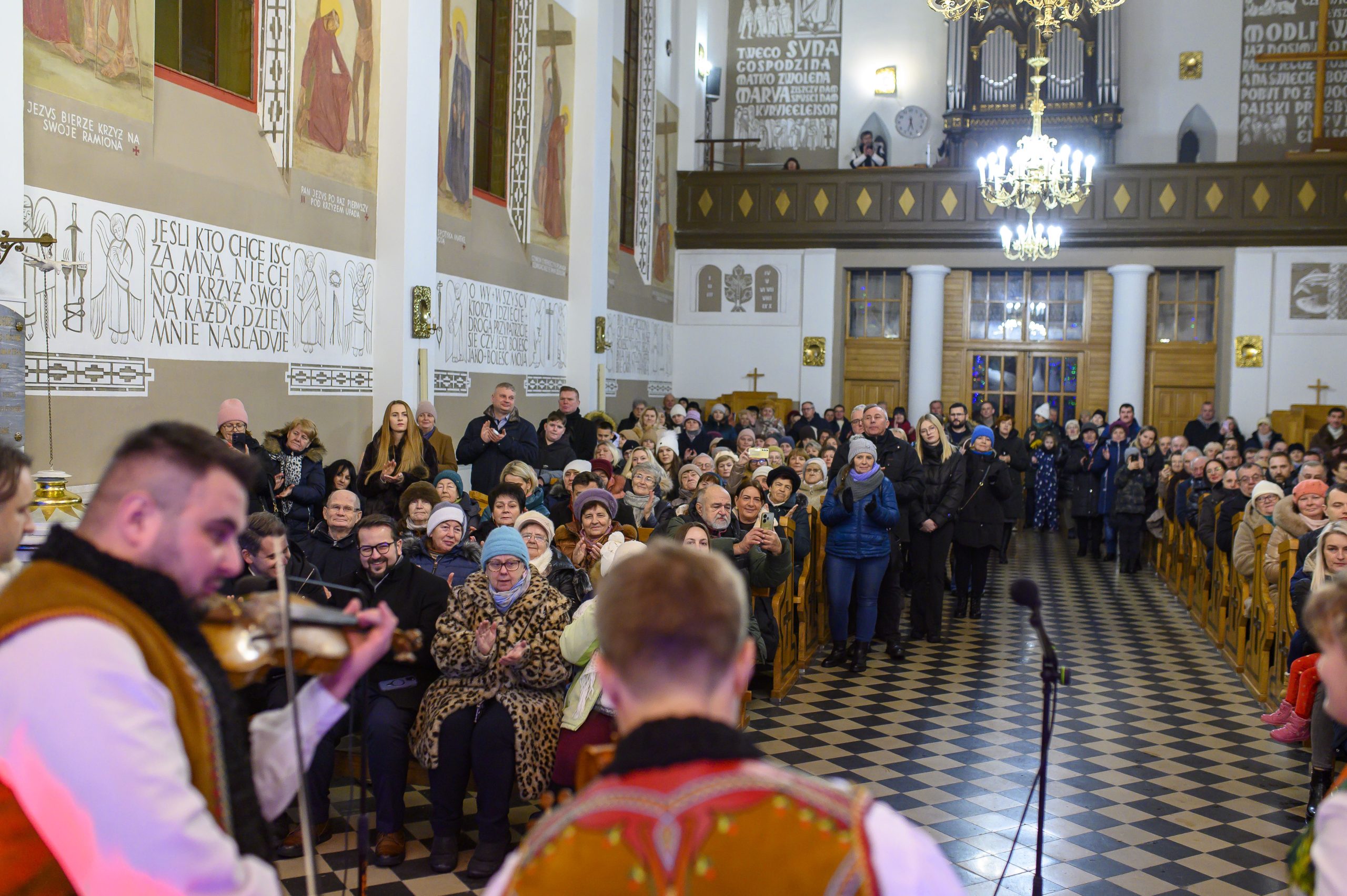 Koncert w kościele, muzycy w tradycyjnych strojach góralskich na pierwszym planie. Publiczność siedzi i stoi, aplauz. Wnętrze zdobione freskami, jasne oświetlenie.