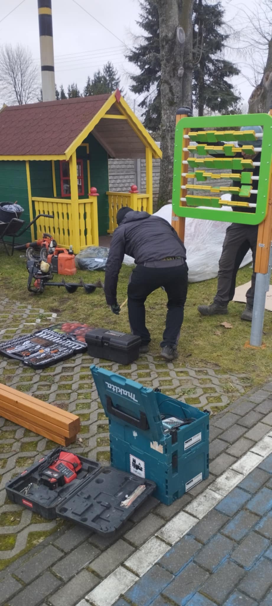 Dwóch mężczyzn pracuje przy placu zabaw z kolorowym domkiem. Obok narzędzia na kostce brukowej. W tle drzewa i szare niebo.