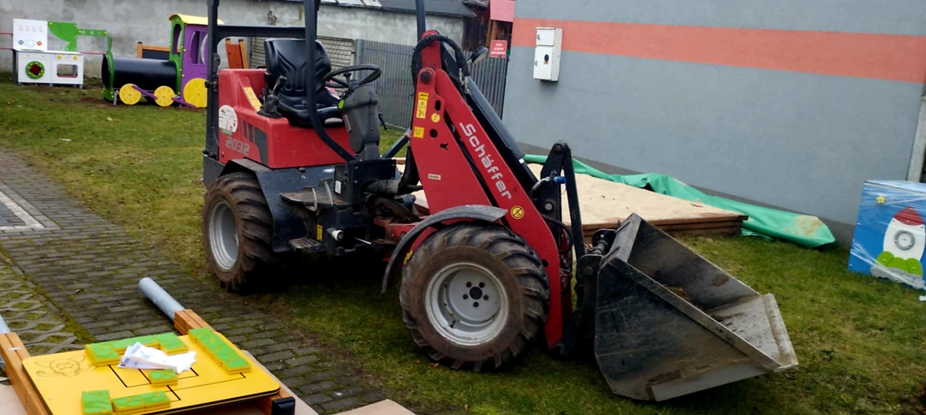 Na trawniku stoi czerwona ładowarka z dużą łyżką. W tle kolorowy plac zabaw i szare ogrodzenie. Na ziemi leży drewniana platforma z zielonymi elementami.