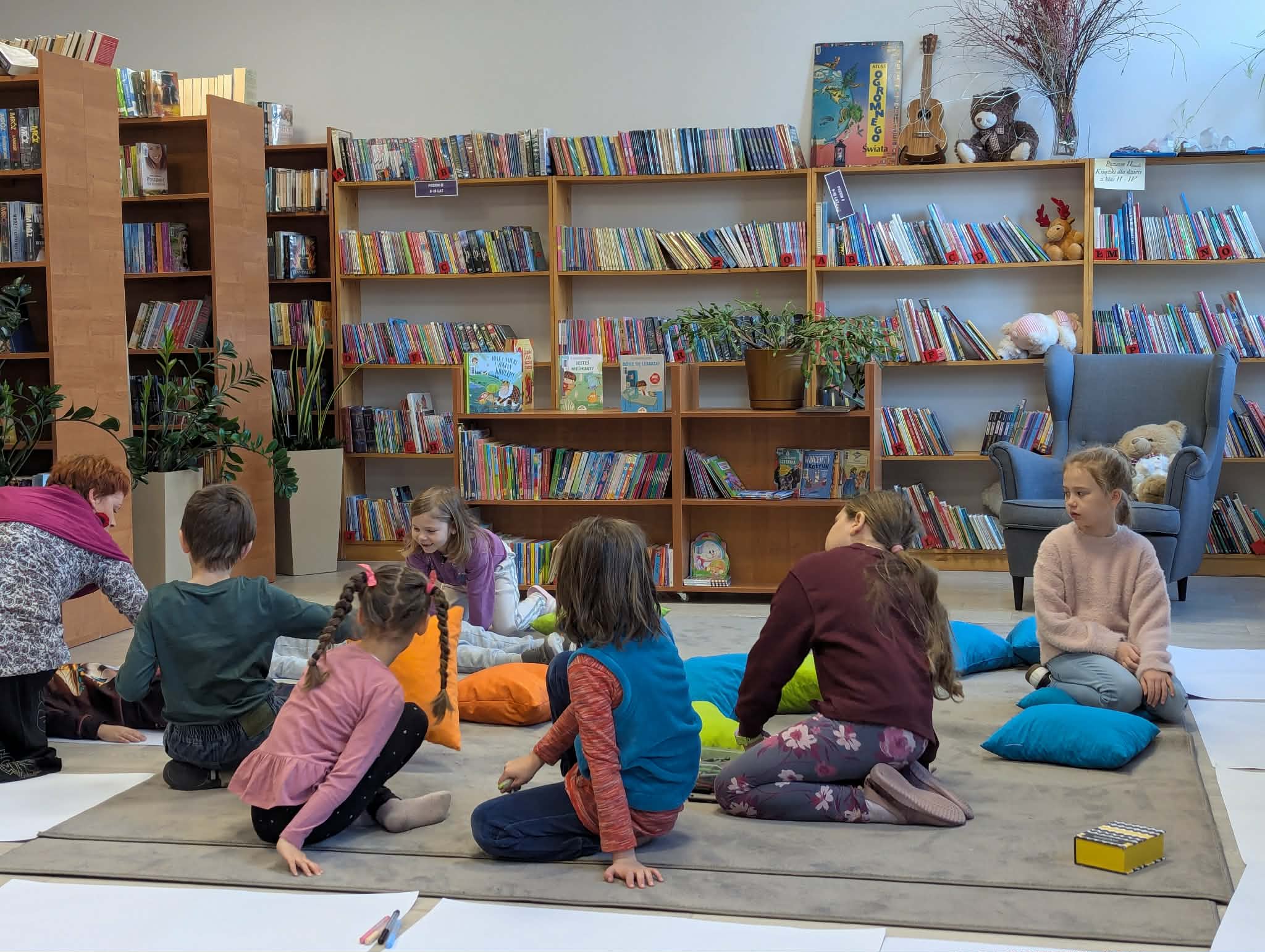 Grupa dzieci siedzi na dywanie w bibliotece, otoczona kolorowymi poduszkami. W tle są półki pełne książek. Jasne ściany i rośliny tworzą przytulną atmosferę.