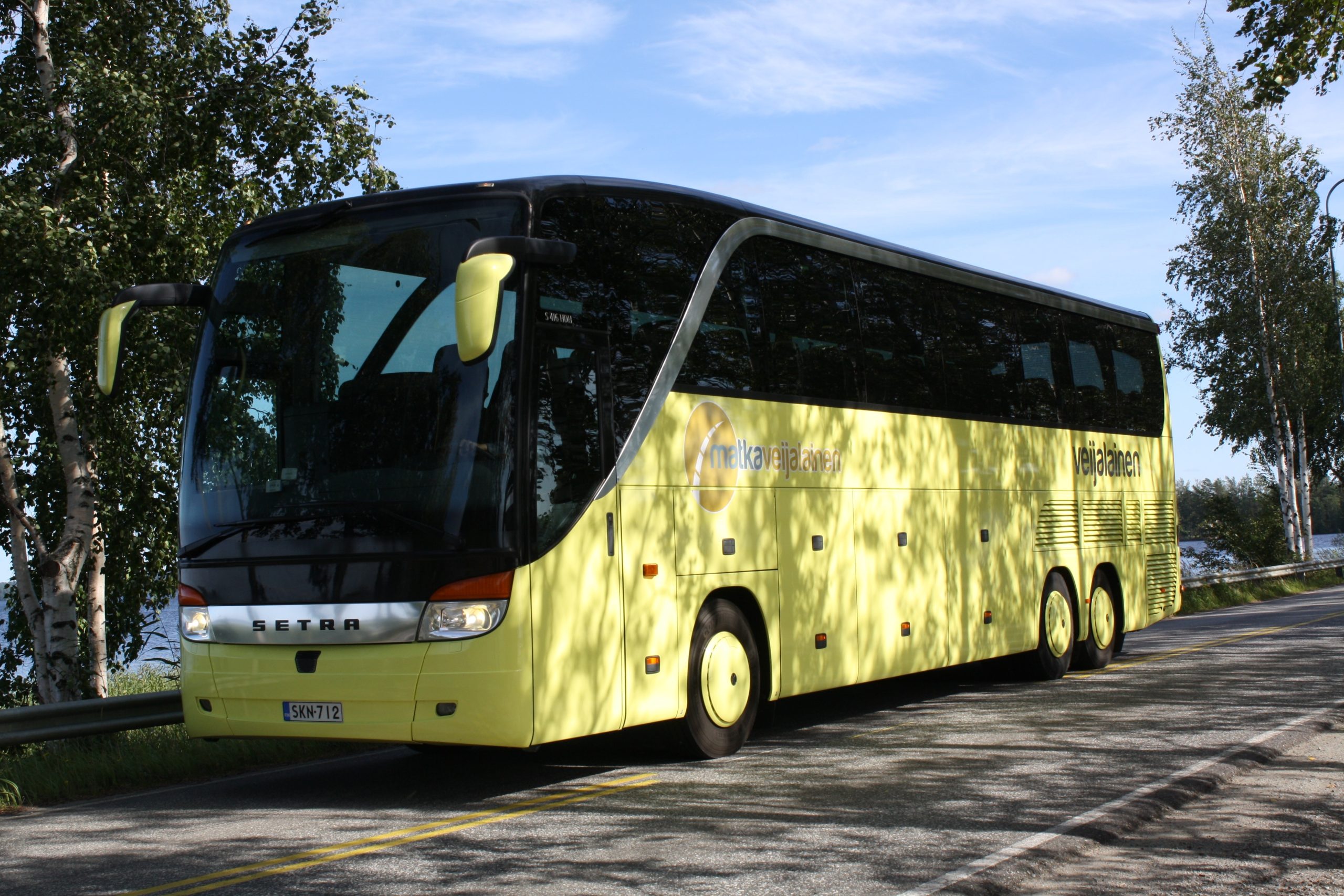 Jasnożółty autobus na drodze z drzewami w tle. Niebieskie niebo i cienie liści na pojeździe. Znaki firmowe na boku.