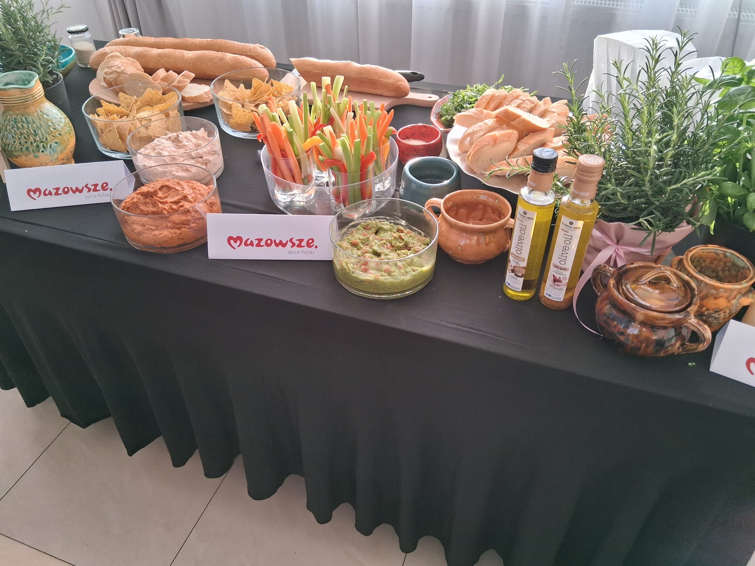 Stół z czarnym obrusem pełen przekąsek: pieczywa, nachosów, warzyw, sosów oraz oliwy. Obok zioła w doniczkach. Tło w jasnych kolorach.