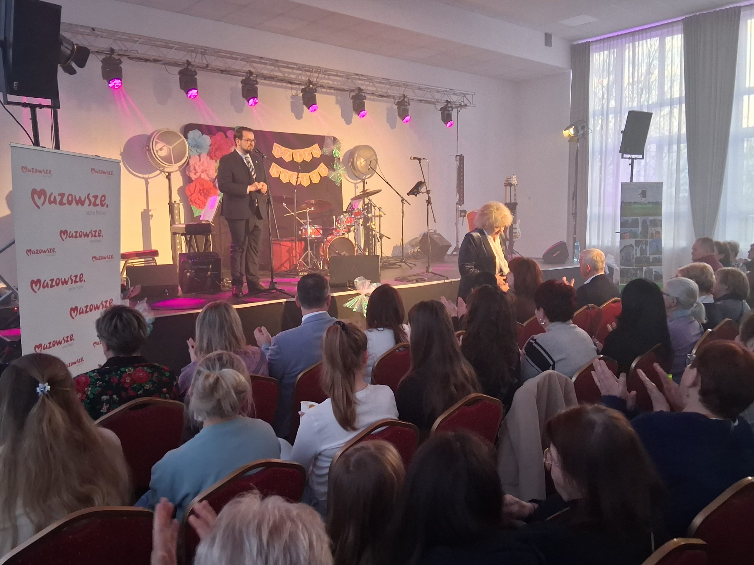 Na scenie jasne oświetlenie, stojący mężczyzna w garniturze, dekoracje z kwiatów. Publiczność siedzi na czerwonych krzesłach. Obok sceny biały baner z napisem „Mazowsze”.
