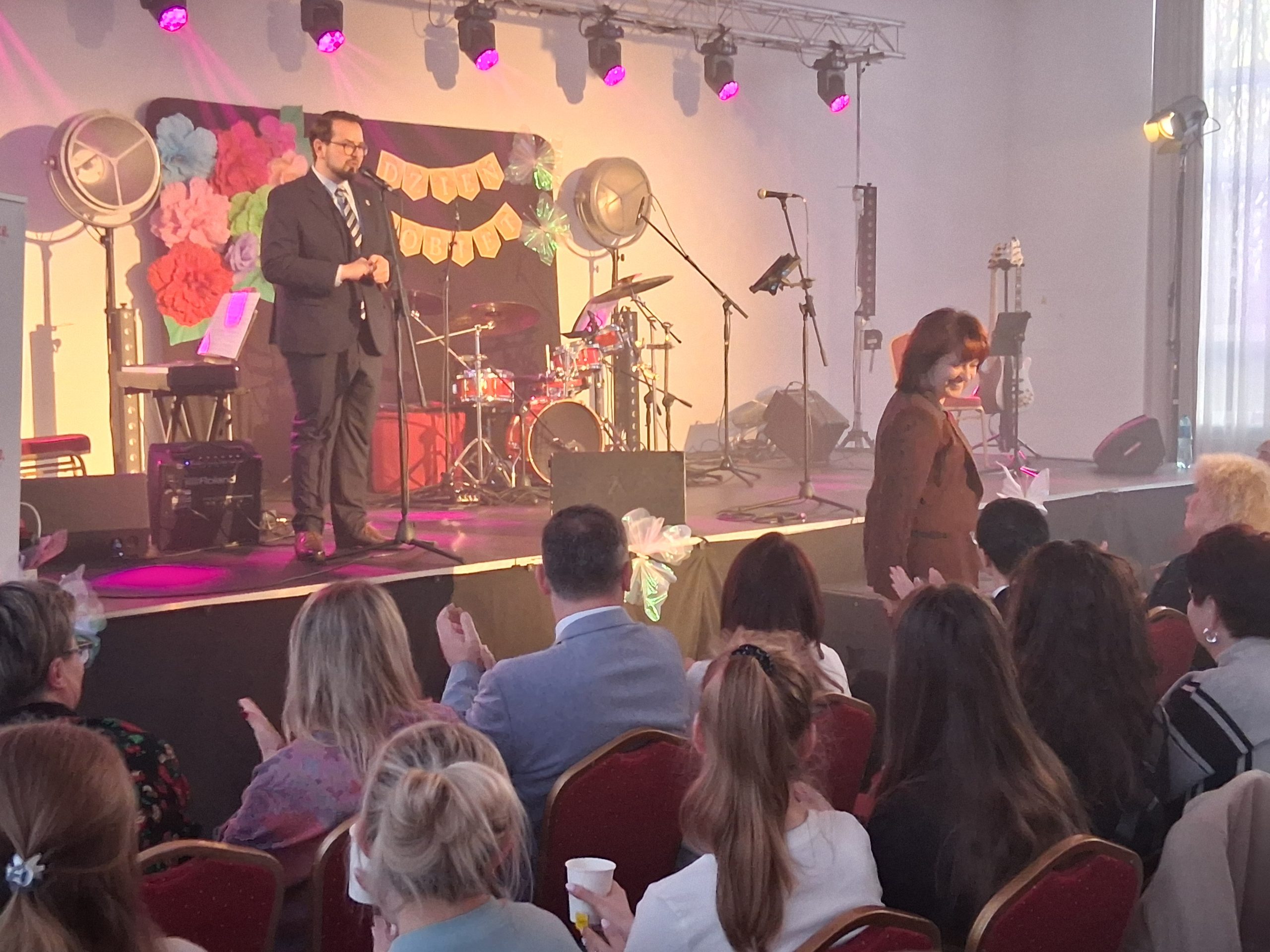 Osoba na scenie przemawia do publiczności. Tło zdobią kolorowe dekoracje i instrumenty muzyczne. W pomieszczeniu panuje przytulna atmosfera.