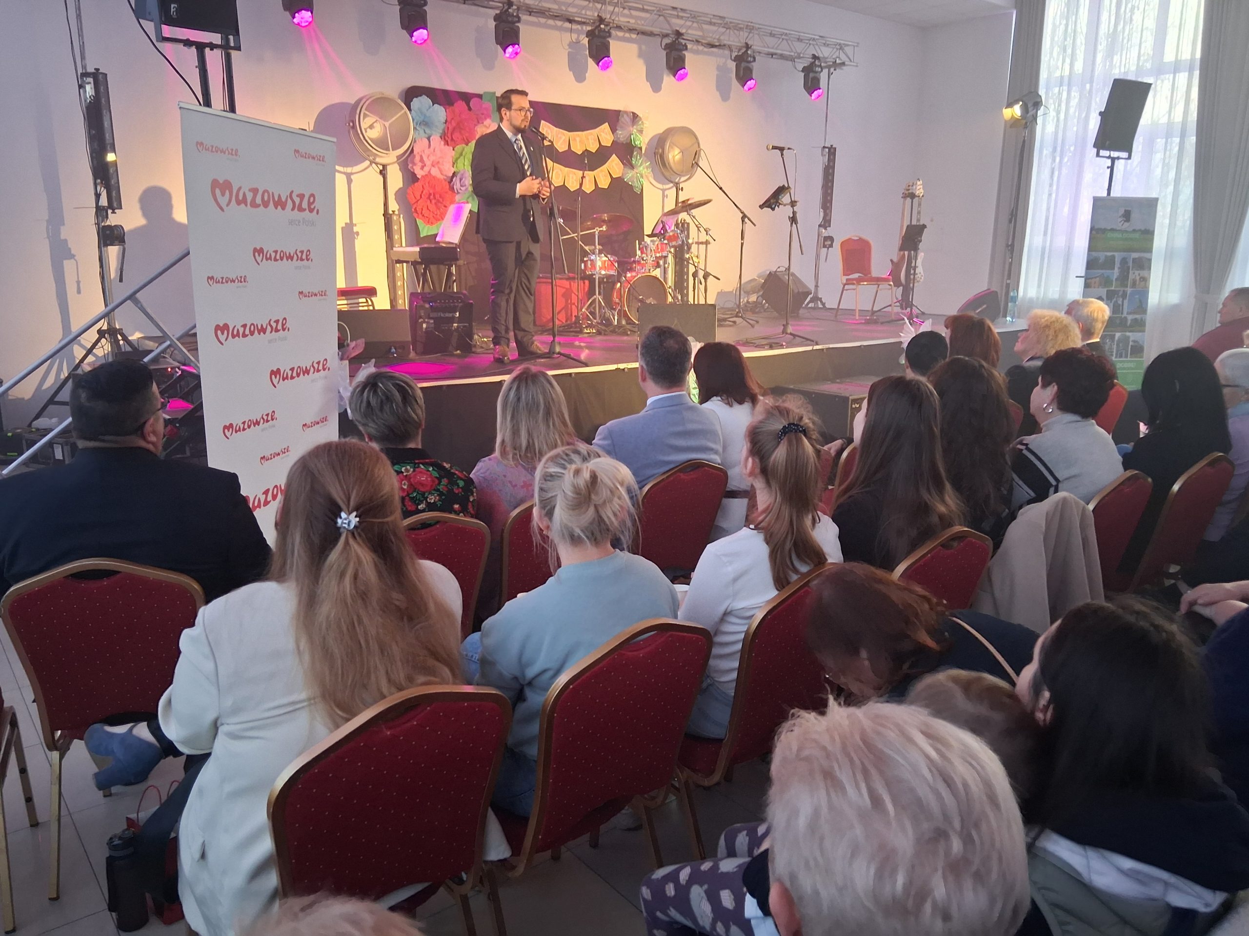Mężczyzna na scenie w ciemnym garniturze przemawia do publiczności. Scena udekorowana kolorowymi kwiatami. W tle widoczne instrumenty muzyczne, a publiczność siedzi na czerwonych krzesłach.