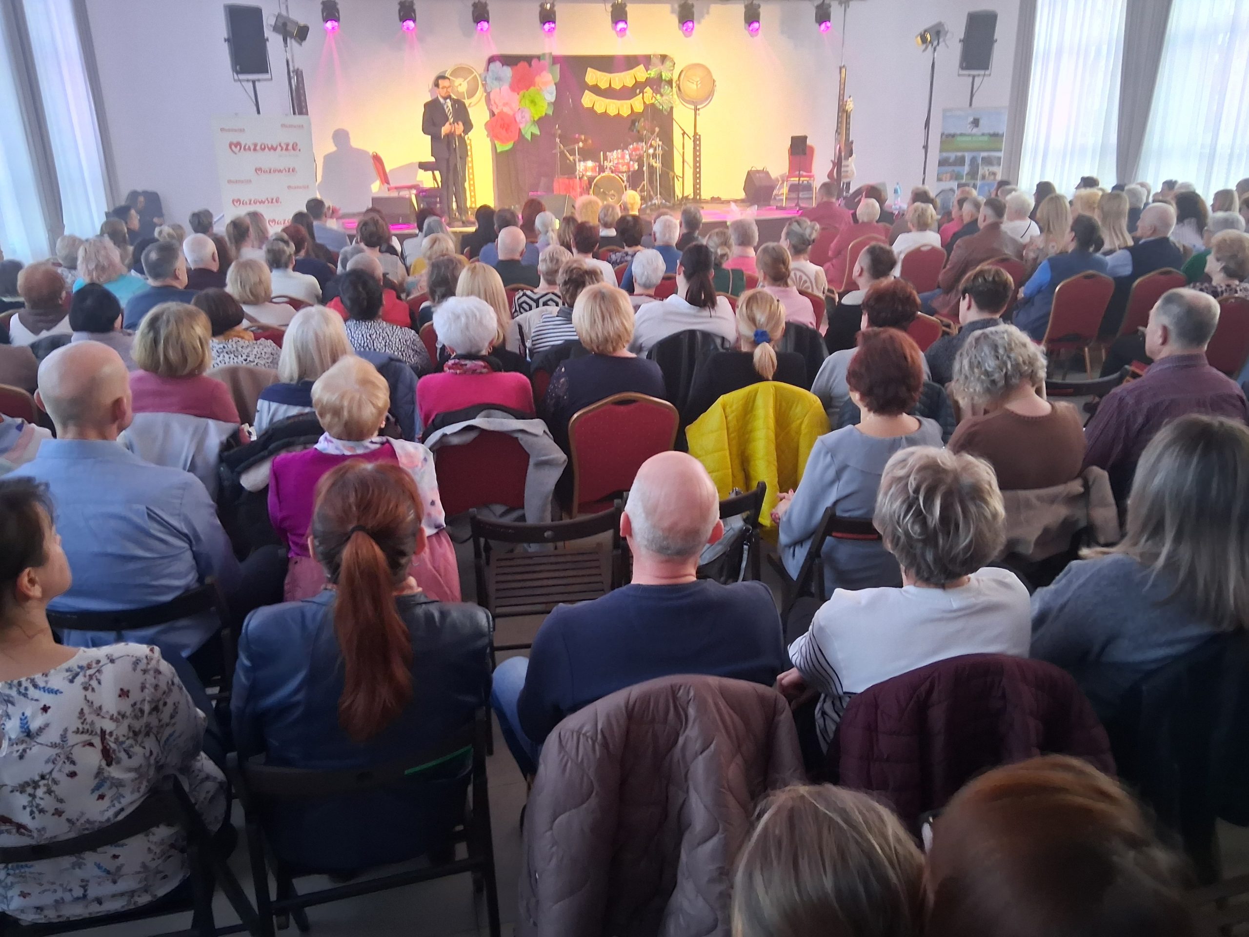 Sala pełna ludzi siedzących na krzesłach, zwróconych w stronę sceny. Na scenie mężczyzna przemawia, dekoracje w jasnych kolorach. W tle oświetlenie w fioletowych odcieniach.