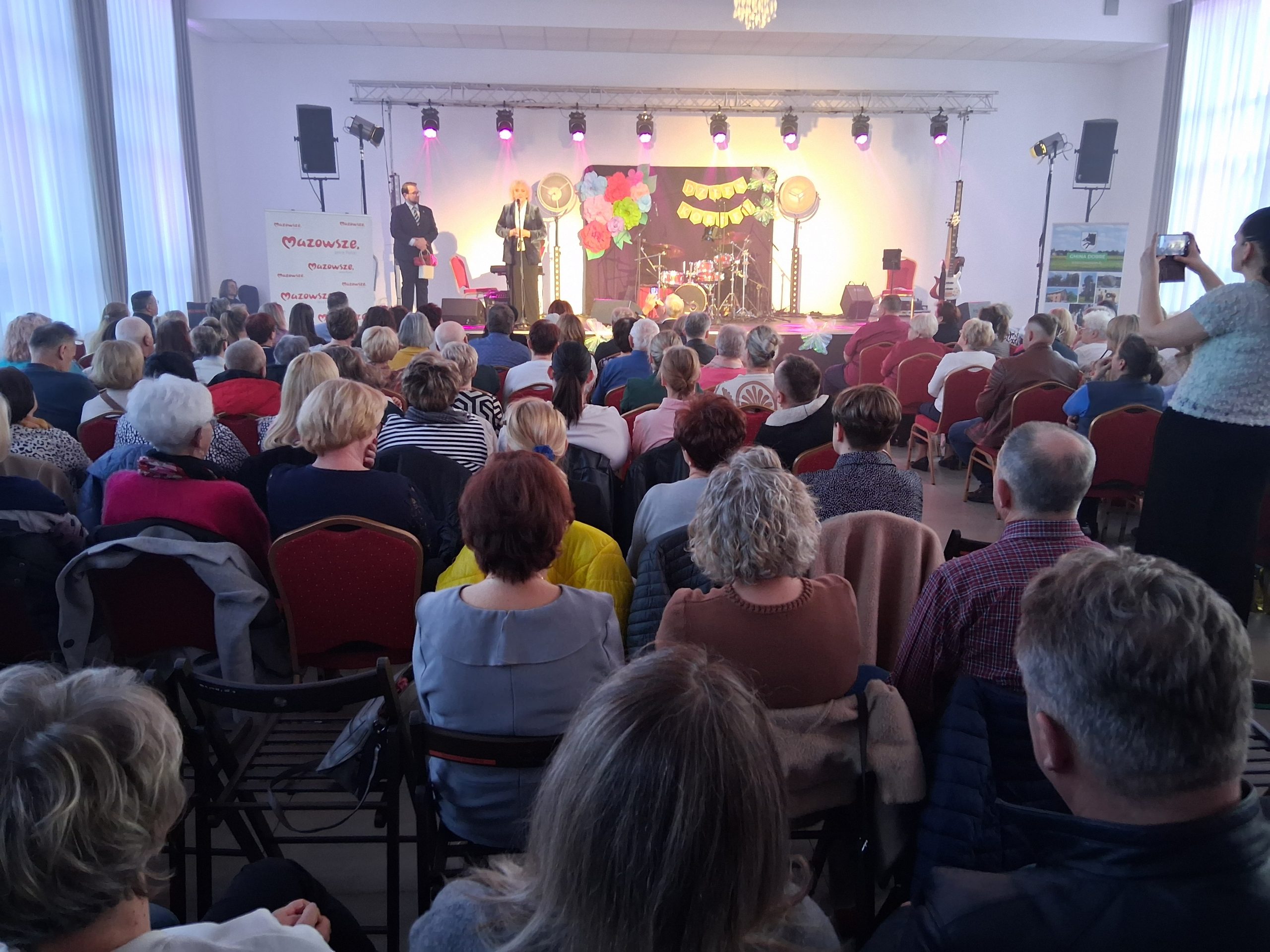 Na zdjęciu widoczna jest sala pełna ludzi zwróconych w stronę sceny. Scena ozdobiona kolorowymi światłami i dekoracjami. Na scenie widoczni są dwaj mówcy.