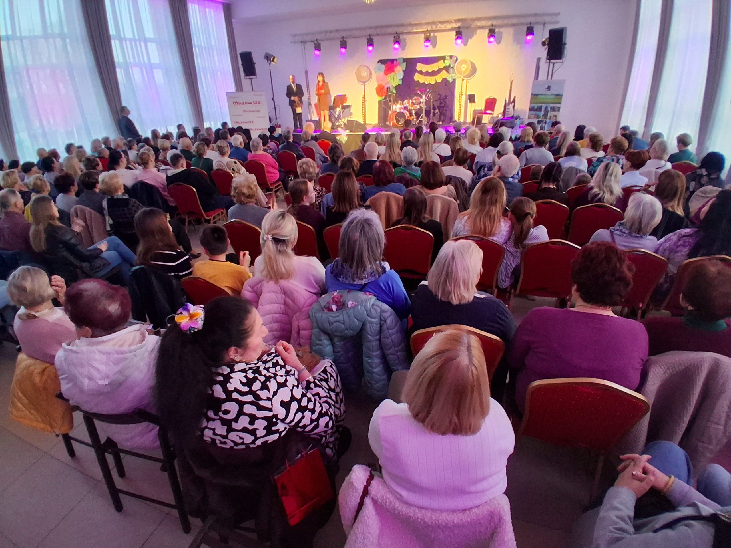 Na zdjęciu jest pełna sala ludzi siedzących na czerwonych krzesłach. Na scenie znajdują się osoby, przy jasnym, kolorowym oświetleniu. Tło zdobią wiszące dekoracje, a za oknami widać zasłony.