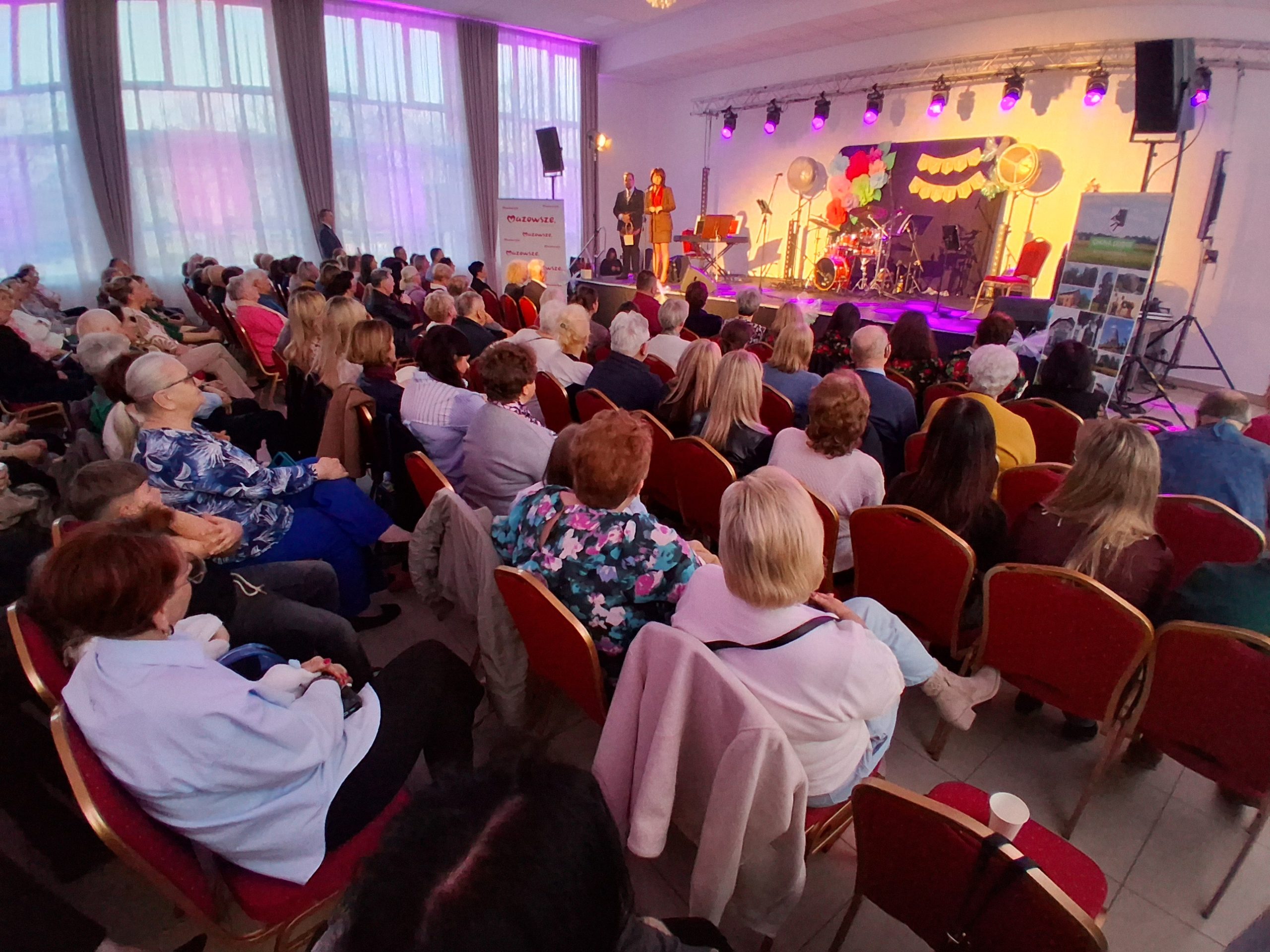 Sala pełna ludzi siedzących na czerwonych krzesłach, scena oświetlona fioletowym światłem. W tle zasłony i stoliki. Na scenie stoi osoba, obok zestaw perkusyjny.