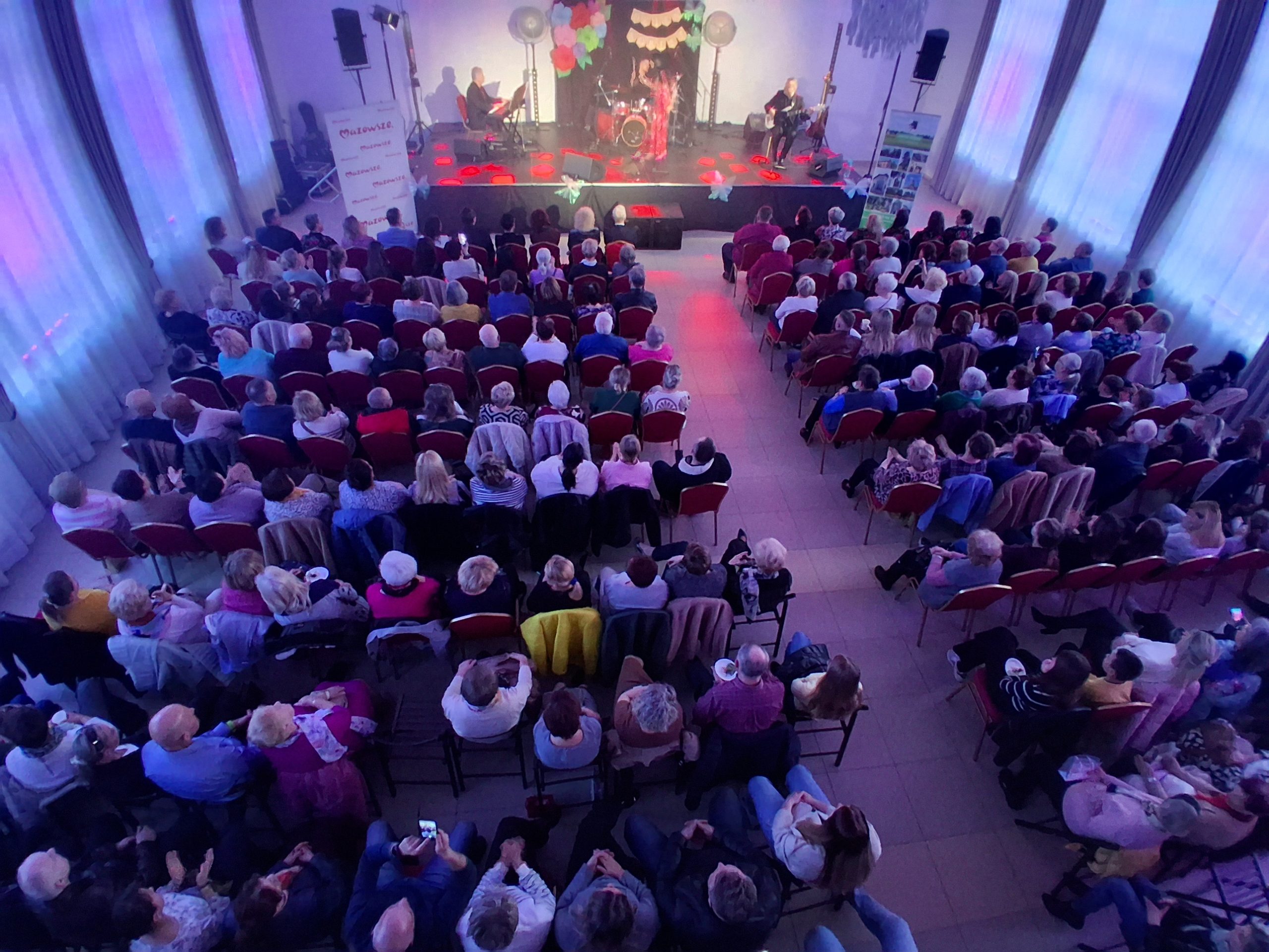 Sala z publicznością siedzącą na czerwonych krzesłach, patrzącą na scenę z zespołem muzycznym. Tło jest jasne, z dużymi oknami po bokach i kolorowymi światłami. Dekoracje na scenie w intensywnych kolorach, atmosfera koncertowa