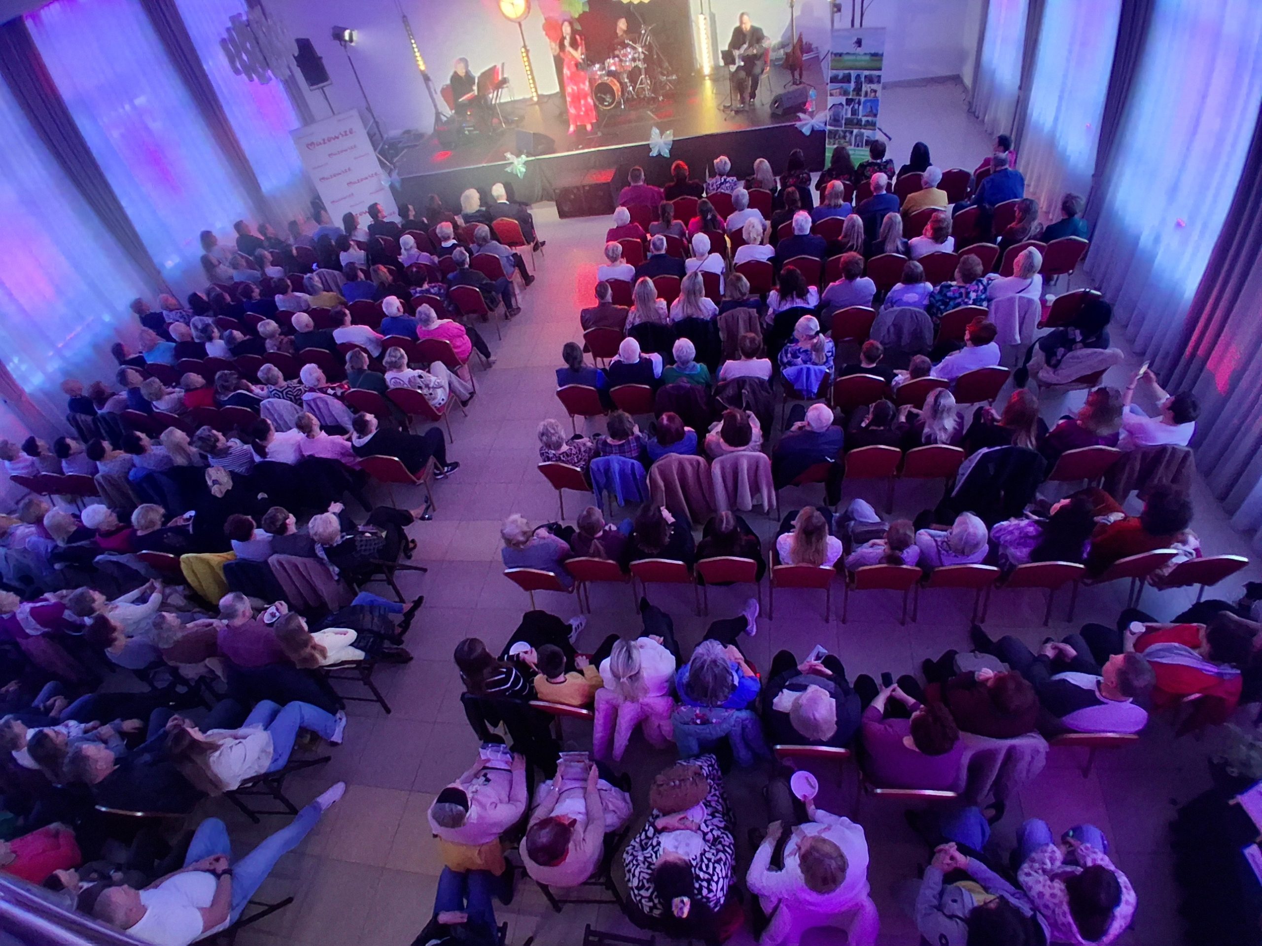 Widok z góry na dużą salę z publicznością skierowaną w stronę sceny. Ludzie siedzą na czerwonych krzesłach w jasnym pomieszczeniu z kolorowymi światłami. Na scenie widać zespół muzyczny.