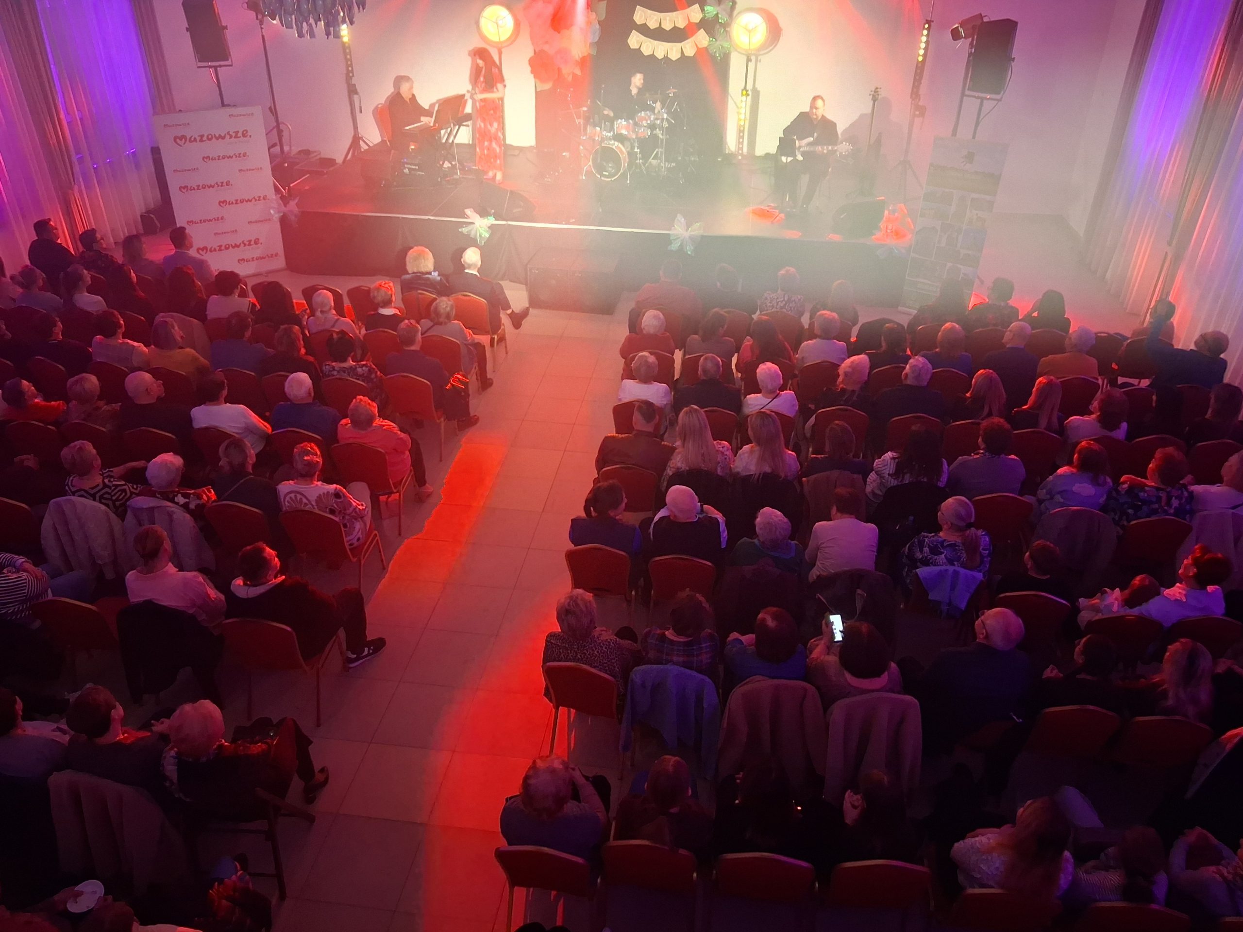 Sala koncertowa w czerwonym oświetleniu. Widownia siedzi przed sceną z muzykami. Tło jest jasne z elementami dekoracyjnymi.