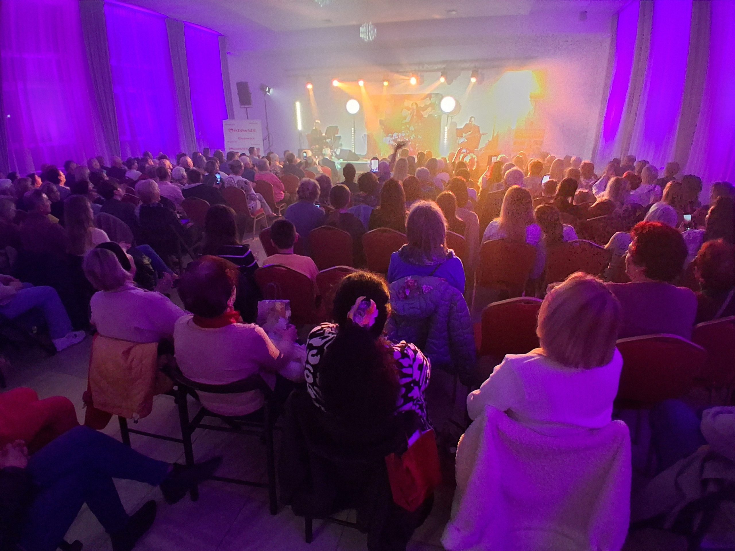 Sala wypełniona ludźmi, scena oświetlona fioletowym i pomarańczowym światłem. Na scenie muzyk. Publiczność siedzi na krzesłach.