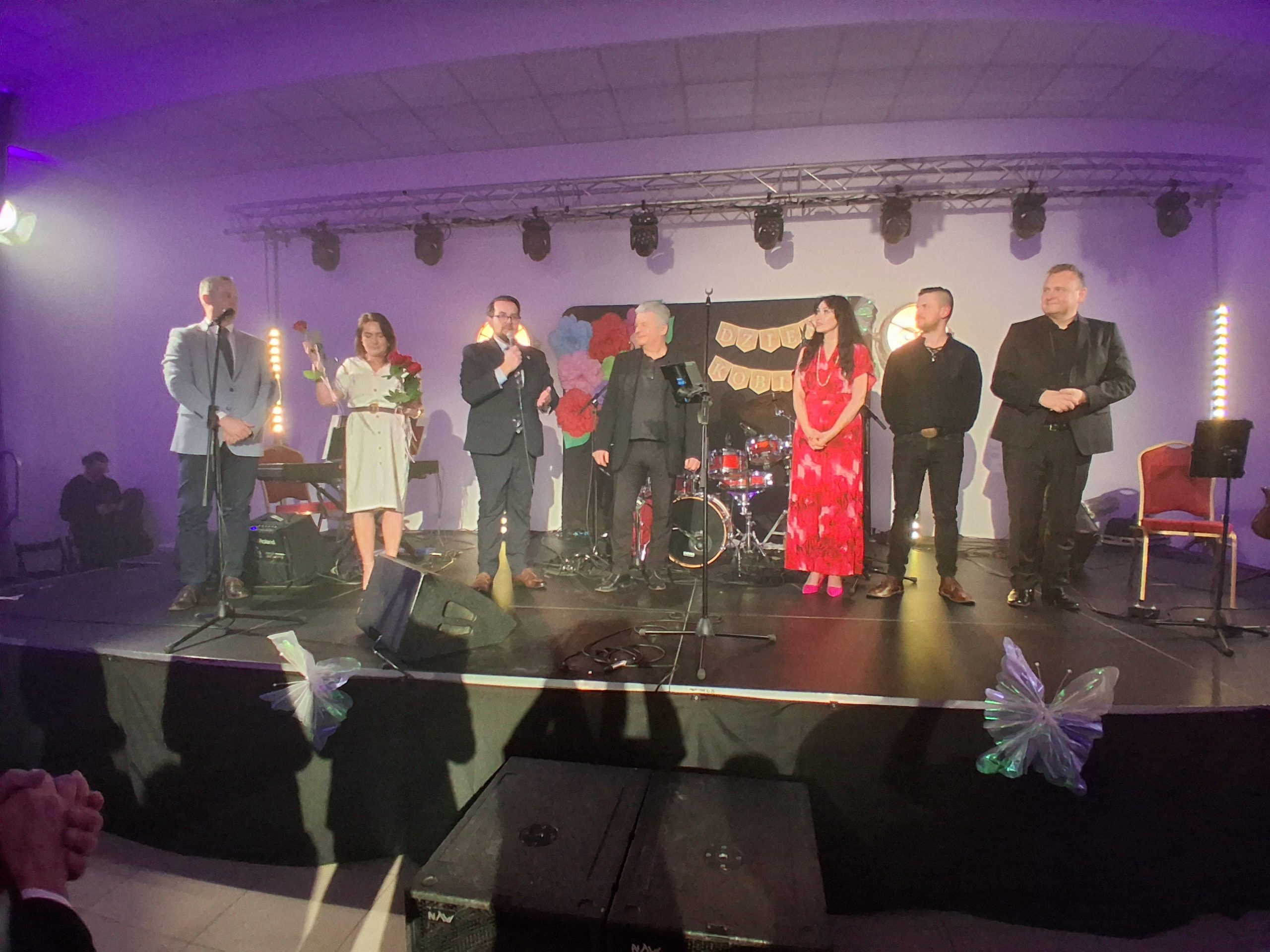 Grupa osób stoi na scenie, część trzyma mikrofony. Tło jest jasne z ozdobą z kolorowych kwiatów. W tle widać instrumenty muzyczne i dekoracje świetlne.