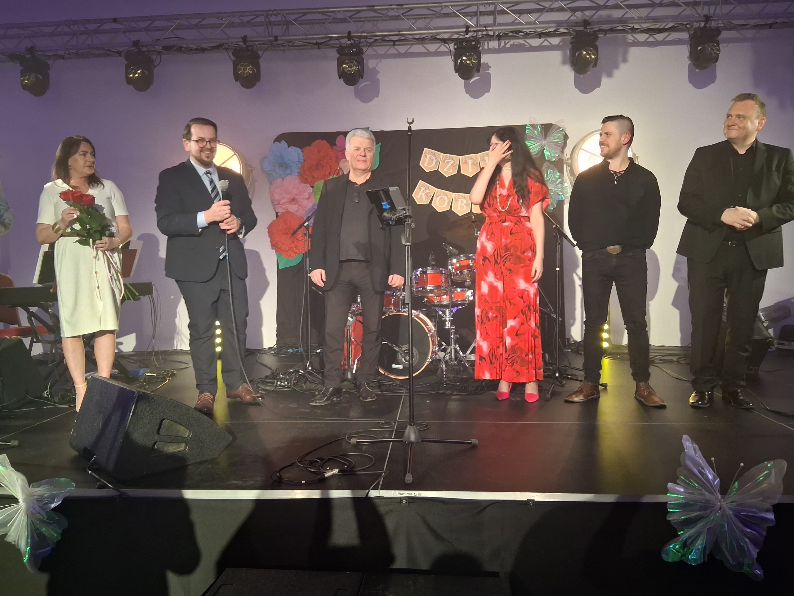 Grupa sześciu osób stoi na scenie, ubranych elegancko. W tle widać perkusję i napis Dzień Kobiet. Dekoracje są kolorowe, dominują czerwone i różowe akcenty.