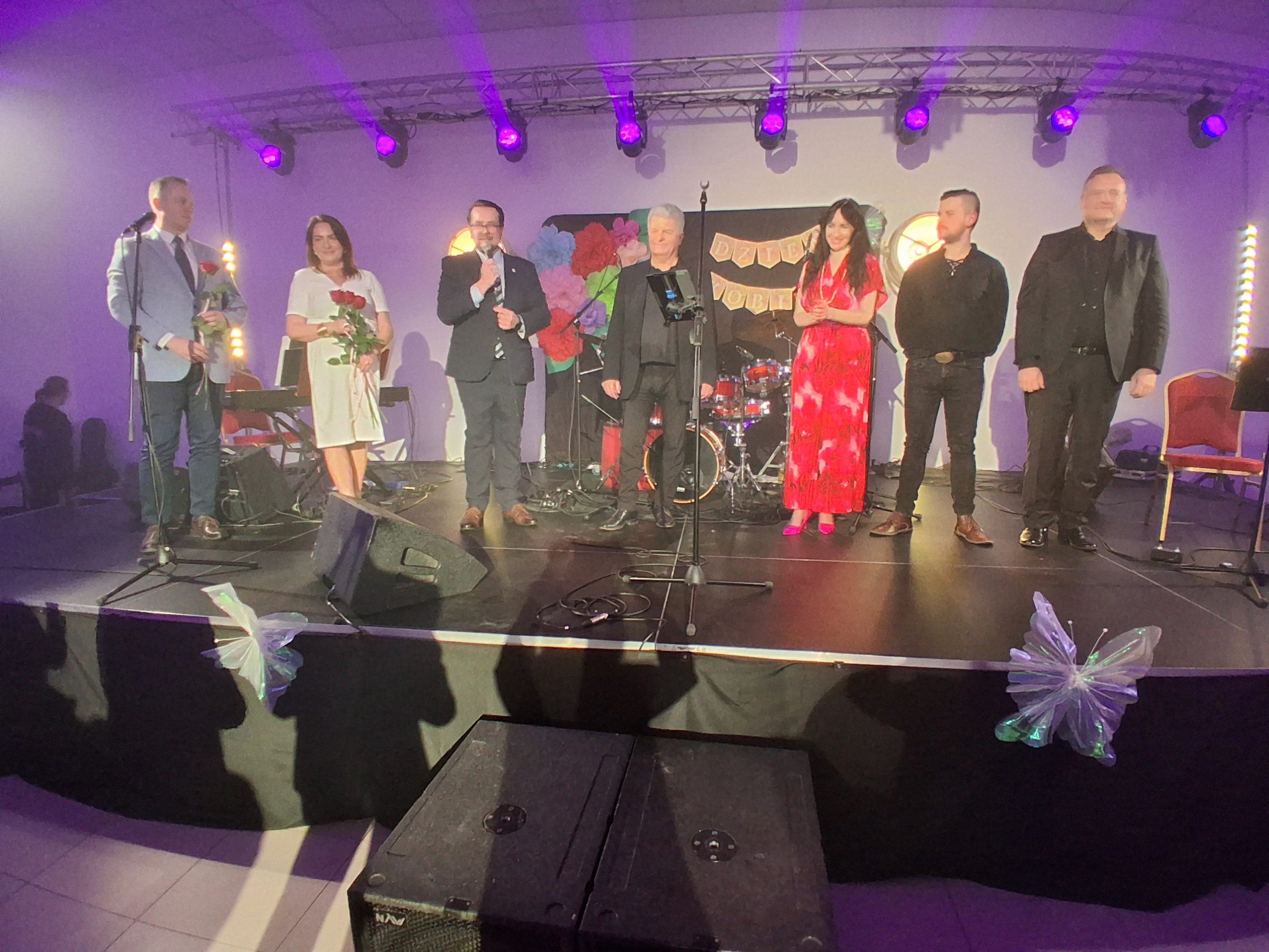 Na scenie stoi siedem osób, w tym kobiety z kwiatami. Tło jest ozdobione napisami i instrumentami muzycznymi. Oświetlenie fioletowe.